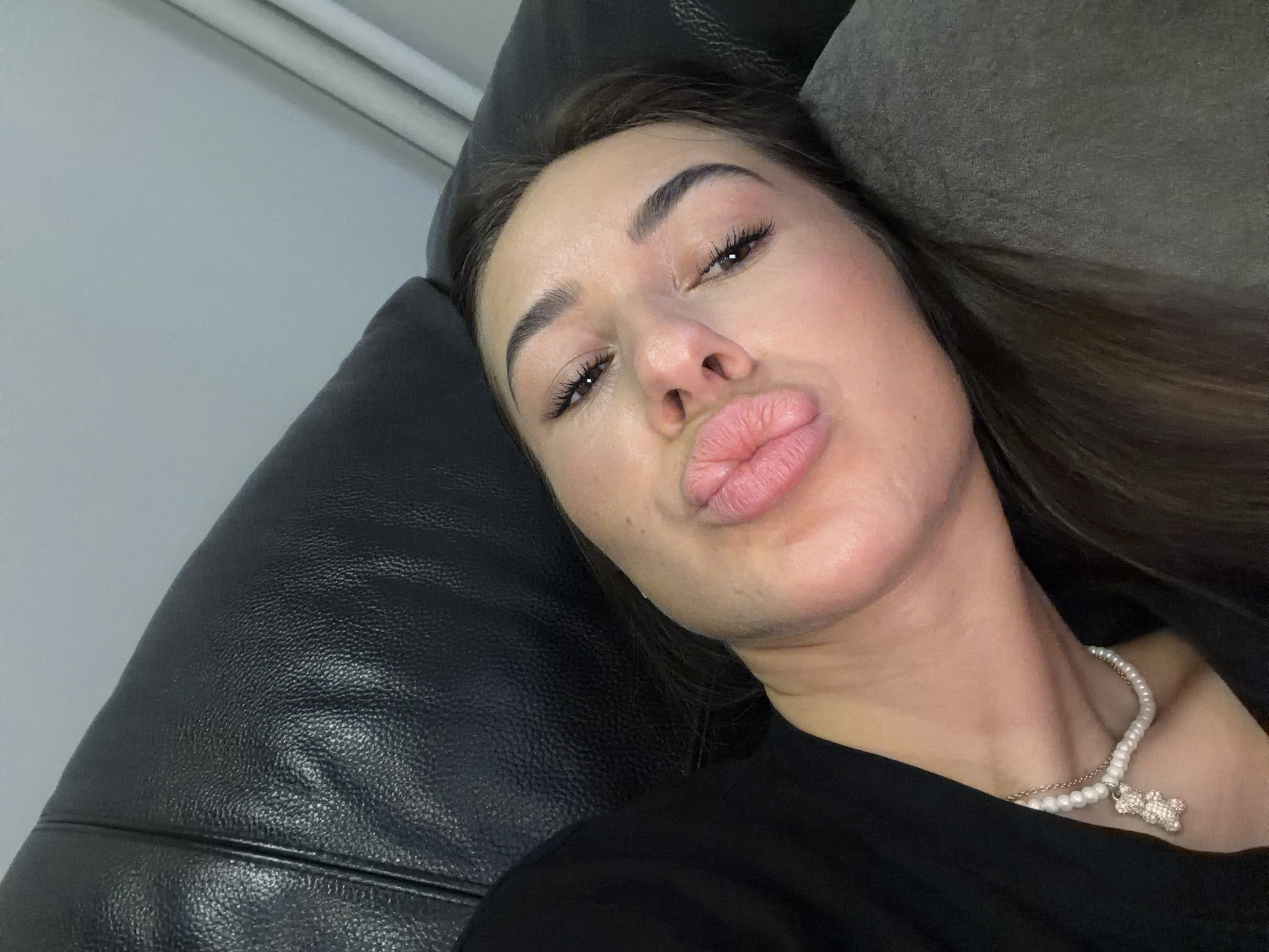 A woman with full lips and dark hair is lying down on a dark sofa, looking at the camera. She is wearing a black top and a pearl necklace with a teddy bear charm. The background is a light-colored wall with vertical blinds. The scene appears to be indoors, likely a home or studio, with no specific city or geographical indicators.