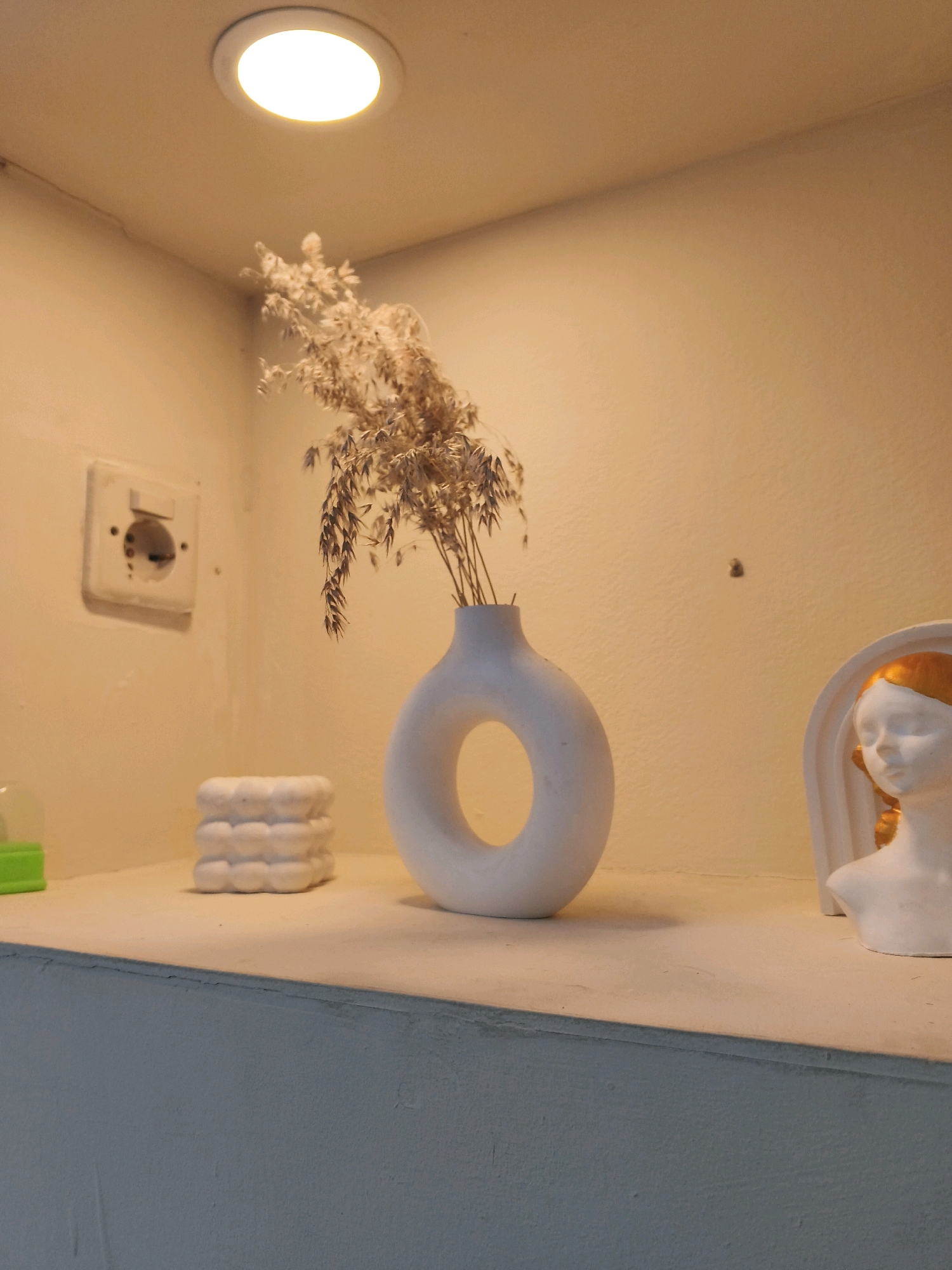 The image shows a shelf with various decorative items, illuminated by a ceiling light. On the left, a white electrical outlet is visible on a light-colored wall. Next to it, a small, white, textured object sits on the shelf. In the center, a white, ring-shaped vase holds dried grass or reeds. To the right, a white bust of a person with gold-colored hair is displayed. The shelf itself is a light color and appears to be part of a wall unit. The background wall is a uniform light color. This scene is not in Ciampel, Indonesia; it is an indoor setting with decorative objects.