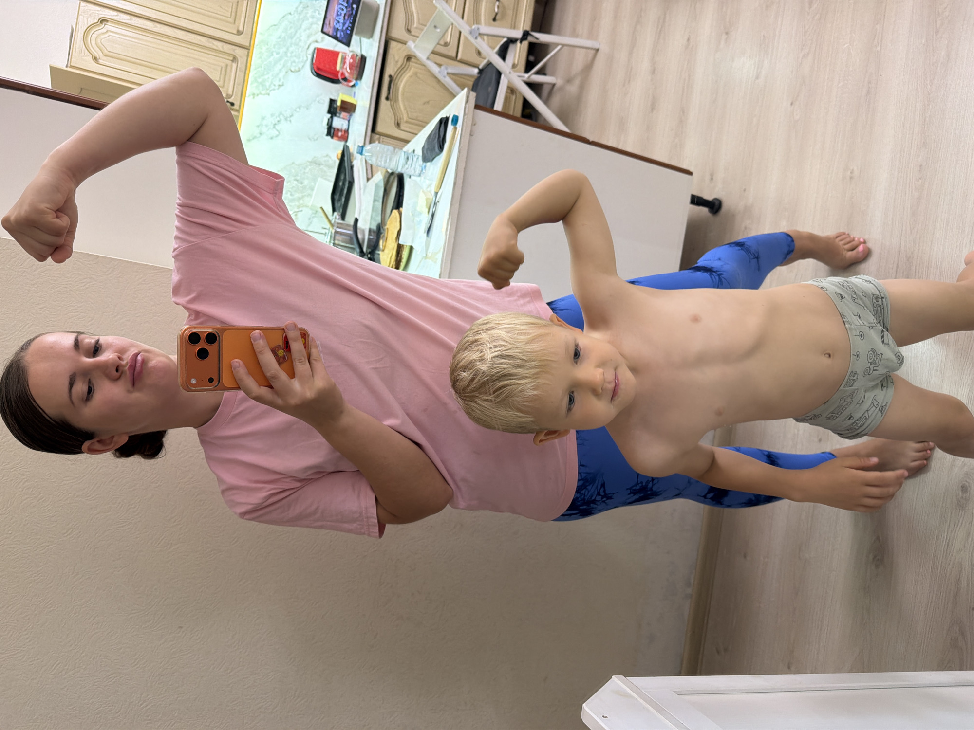 In an indoor scene, a woman and a young boy playfully pose, flexing their biceps. The image appears to be a selfie taken by the woman, holding an orange smartphone in her left hand, with the camera facing a reflective surface or a mirror. The woman, with dark hair pulled back, wears a pink t-shirt and blue patterned leggings. The young boy, with blonde hair and blue eyes, is shirtless, wearing light grey patterned underwear and blue leggings matching the woman's. He stands beside the woman, mirroring her pose.
The background shows a domestic setting, likely a kitchen or a room with kitchen elements. On the right, there's a light-colored wooden floor. Behind the subjects, a kitchen counter or table holds various items, including a white bottle, a red container, and a tablet displaying the time "10:13". Wooden cabinets are visible above the counter, and a white folding chair is partially seen. The walls are a neutral, light color. The overall atmosphere is lighthearted and familial, captured in what appears to be a home in Sochi, Russia.