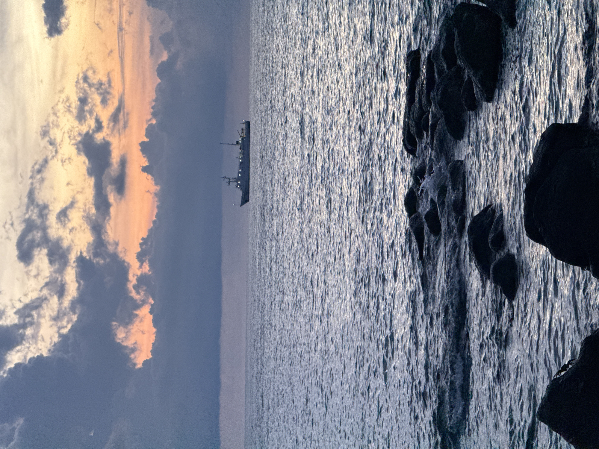 This is a photograph of the sea near Lý Sơn Island, Vietnam, at dusk. The sky is filled with dramatic clouds, tinged with orange and pink from the setting sun, contrasted with darker, ominous gray clouds. The sea is choppy, with visible waves and whitecaps, and reflects the colors of the sky. In the distance, a ship sails on the horizon, silhouetted against the colorful sky. In the foreground, dark, rough rocks are scattered along the shoreline, some partially submerged in the water. The overall mood of the image is serene yet dramatic, capturing the raw beauty of nature at the edge of day.