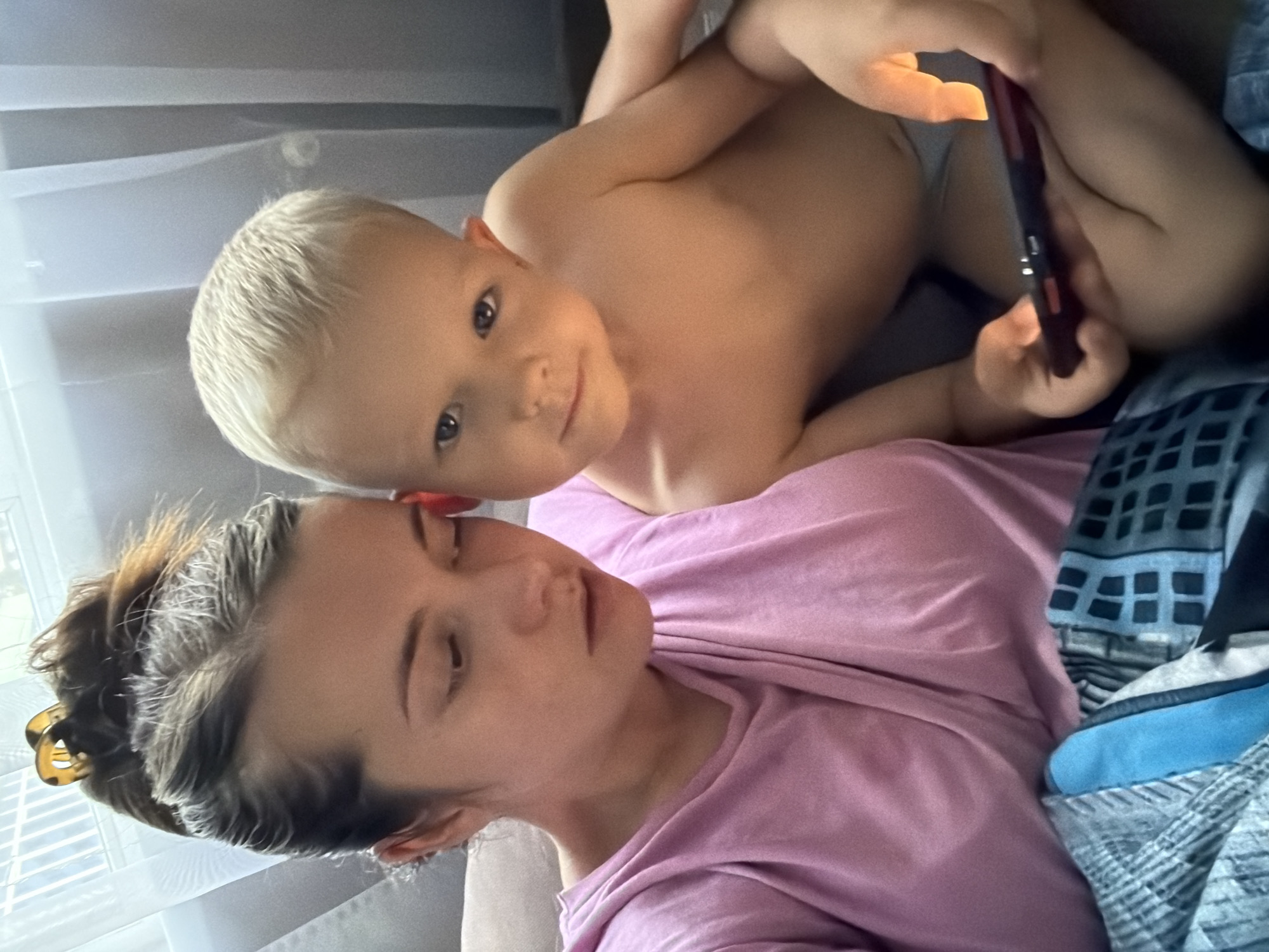 This intimate indoor photograph, set in Sochi, Russia, features an adult female and a young child in a relaxed, close-up moment. The blonde-haired child, appearing to be a toddler or preschooler, lies on top of the adult, looking directly at the camera with a contented, soft smile. The child is shirtless or wearing light underwear and holds a dark smartphone with both hands.
The adult, likely resting, is seen beneath the child, wearing a pink t-shirt. Her dark hair is pulled back with a tortoiseshell claw clip, and her eyes are closed or nearly closed, conveying a sense of peace and relaxation. Her face shows a serene expression.
The setting appears to be a bedroom or living area, with soft daylight illuminating the scene through sheer curtains in the background. A patterned duvet or blanket in shades of blue, grey, and black is visible on the right side of the frame. The image captures a quiet, tender daytime moment between the two subjects, emphasizing their closeness. There is no visible text in the image.