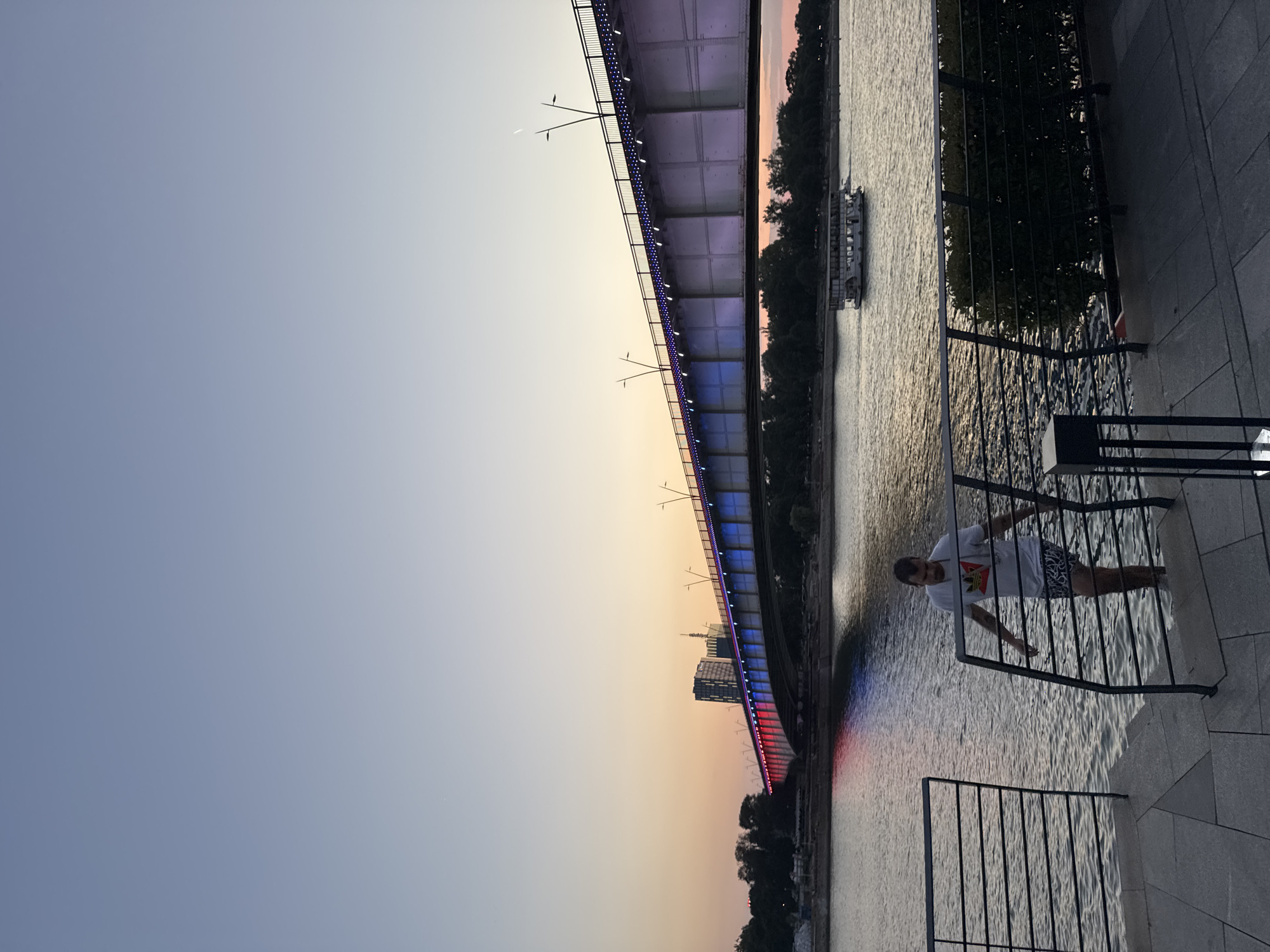 The image depicts a sunset scene along the Sava River in Belgrade, Serbia. A man, wearing a white t-shirt and patterned shorts, is partially submerged in the river, holding onto a railing. He is standing on stone steps leading into the water. In the background, a large, illuminated bridge spans the river, displaying red and blue lights. The bridge is lined with streetlights and smaller lights. A tour boat is visible on the river in the middle ground. Trees and buildings are present on the far bank of the river. The sky exhibits a gradient of colors from pale blue at the top to orange and pink hues near the horizon. The foreground features a paved walkway with railings and some greenery.