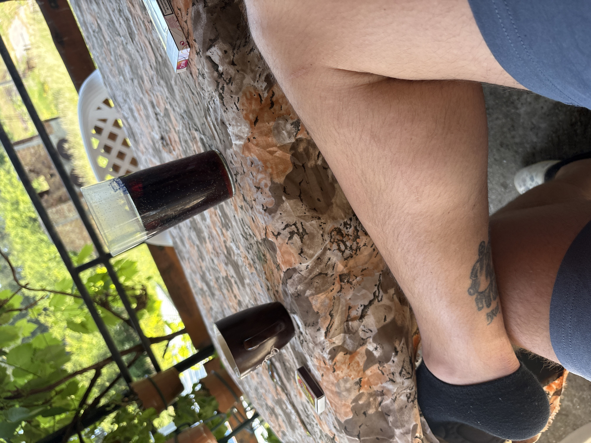 The image presents a close-up, angled view of a person's lower body and a patterned table in an outdoor or semi-outdoor setting. The perspective is from above, looking down.
In the center and right, the bare, slightly hairy legs of an individual wearing blue shorts are visible. On the lower part of the right leg, above a black ankle sock, there is a dark tattoo featuring an abstract design or figure with some illegible script underneath. The left foot is also wearing a black ankle sock.
The table surface, which occupies the left and central areas, has a distinct mottled pattern resembling marble or granite, with shades of beige, brown, and black. On the table, a clear glass filled with a dark red beverage, possibly wine or a dark soda, is prominently featured. The glass has some condensation and a small, partially visible blue and white label or logo near its rim. Nearby, a dark brown ceramic mug with a handle is placed. Also on the table are two small rectangular items: a matchbox with a red and yellow design, and a white and pink cigarette pack. The cigarette pack clearly displays a health warning: "WARNING: SMOKING KILLS" along with other faint text, likely including the brand name "Rothmans."
The background reveals a vibrant green natural environment, with lush foliage, possibly vines or dense leaves, seen through a dark metal railing. A white plastic garden chair is partially visible behind the table on the left side. The scene is brightly illuminated by natural daylight, suggesting a sunny day. The overall impression is one of leisure and relaxation in a natural, possibly rustic, environment, consistent with a rural location such as Staro Selo, Bulgaria. The activity depicted appears to be casually sitting outdoors, enjoying a drink and possibly smoking.