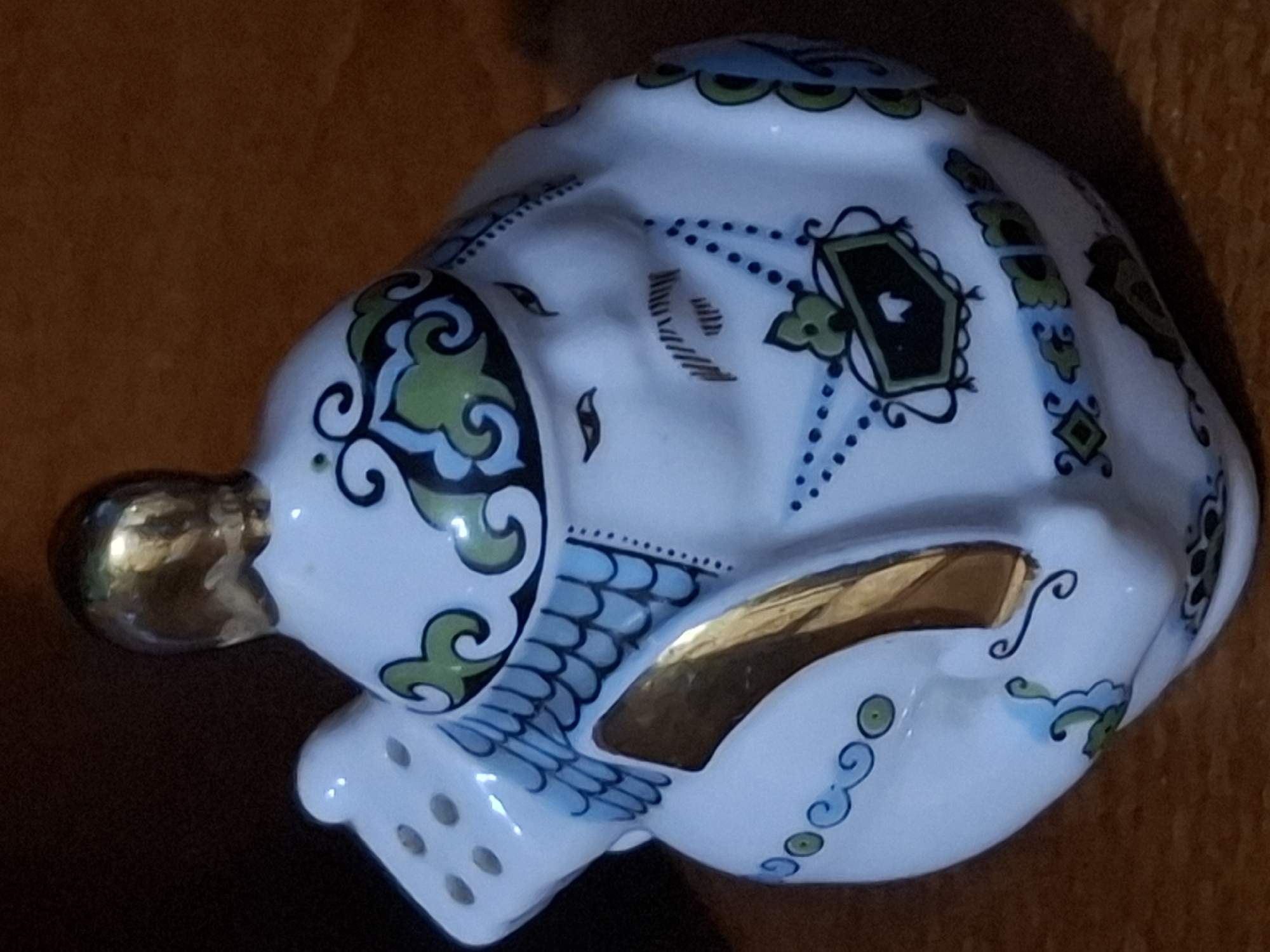 This is a close-up, slightly angled, eye-level shot of a small, white, ceramic teapot or figurine. The object is adorned with blue and green decorative patterns, and has gold accents on its lid and a large, vertical strip on its side. The figurine appears to be shaped like a stylized face with a mustache and eyes painted on. It is resting on a wooden surface, which is visible in the foreground and background. The lighting is dim, creating shadows and highlighting the glossy finish of the ceramic. There is no indication of the city of Almaty, Kazakhstan, or any specific urban environment.