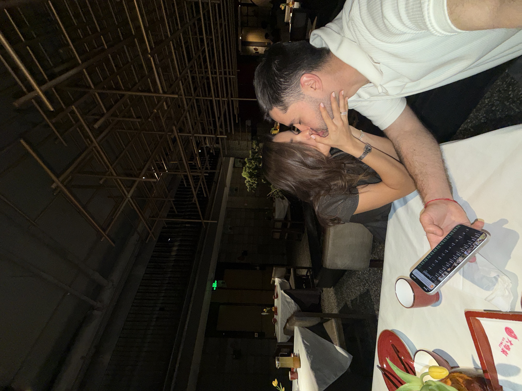 A couple is kissing at a table in a restaurant in Dongcheng District, China. The man is wearing a white shirt. The woman has her hand on his face and is wearing a watch. A phone is being held in the man's hand. A white tablecloth is on the table with plates and food. There are other tables and chairs nearby, and a bamboo structure is visible above.