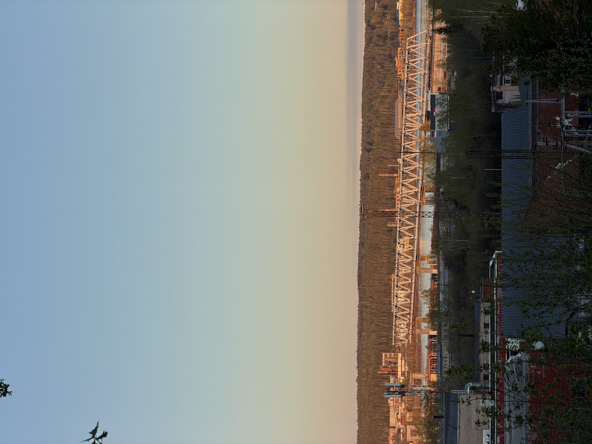 The image shows a view of Perm, Russia, possibly taken from a high vantage point. A large metal bridge spans a river, possibly the Kama River, with dense forest along the riverbank. In the foreground, buildings and industrial structures are visible, including brick buildings with metallic roofs and overhead power lines. The sky is a gradient of blue and yellow, indicating sunrise or sunset. The image is rotated 90 degrees counter-clockwise.