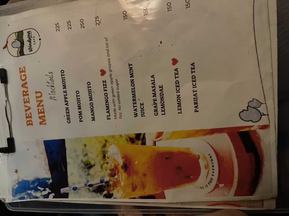 This media file displays a beverage menu from "Windsong CAFE," likely in Dibrugarh, India. The image is taken from a slightly angled, top-down perspective, showing a laminated or plastic-sleeved menu.
The main subject is the menu itself, which lists various drinks and their prices. The menu is divided into sections, with "BEVERAGE MENU" prominently displayed, followed by "Mocktails." A logo featuring a cloud-like shape and the text "Windsong CAFE" is visible at the top.
Visible text includes:
* **Mocktails:**
* GREEN APPLE MOJITO: 225
* POM MOJITO: 225
* MANGO MOJITO: 250
* FLAMINGO FIZZ: 275, with a description: "Made with green apple grapes and lot of fizz. No added sugar." This item, along with "LEMON ICED TEA," has a small red heart icon next to it, possibly indicating a recommended item.
* **Other beverages:**
* WATERMELON MINT JUICE: 150
* GRAPE MASALA LEMONDAE: 150
* LEMON ICED TEA: 150
* PARIJAT ICED TEA: 150
Prices are listed in what appears to be Indian Rupees.
At the bottom of the menu, there is a visual of a vibrant reddish-orange drink in a glass with a blue and white striped straw, suggesting a refreshing beverage. Partial text, "LONG IS FOR EVERYDAY," is visible around the base of the glass in the image. The general activity depicted is someone reviewing a cafe menu. The lighting suggests an indoor setting, possibly during the daytime, but no specific time or weather can be definitively determined.