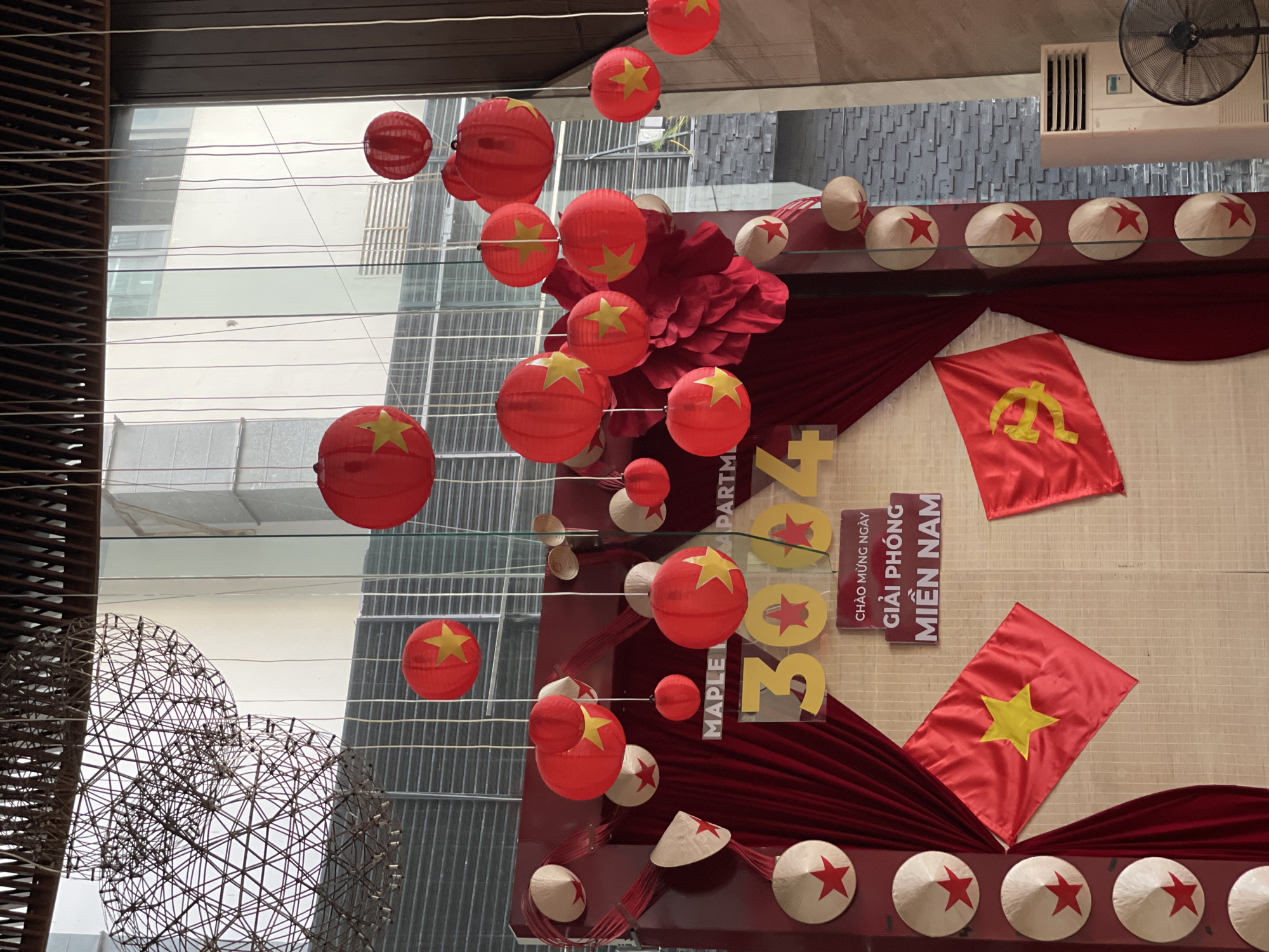 This image captures an indoor space adorned with festive decorations celebrating a Vietnamese national event. The predominant colors are red and yellow, reflecting the national flag of Vietnam. Numerous red paper lanterns, each featuring a yellow five-pointed star, hang at various heights, creating a vibrant overhead display.
Below the lanterns, traditional Vietnamese conical hats (nón lá) are arranged in a row, each decorated with a red star, resting on a red ledge. A backdrop of draped red fabric frames two flags: one is the national flag of Vietnam (red with a yellow star), and the other is a red flag bearing a yellow hammer and sickle emblem. Large yellow numbers "2024" are prominently displayed, indicating the year of the celebration. Next to these numbers, a vertical sign reads "CHÀO MỪNG NGÀY GIẢI PHÓNG MIỀN NAM," which translates to "Welcome Liberation Day of the South," commemorating Vietnam's Reunification Day on April 30th. Partial text "MAPLE" and "PARTMENT" are also visible.
Through the glass panels that form the background, other modern buildings can be seen, suggesting an urban environment, consistent with Nha Trang, Vietnam. A large, spherical wire sculpture is visible on the left. The scene evokes a celebratory and patriotic atmosphere, likely in a public building or commercial establishment. A ceiling fan and an air vent are visible at the top right of the frame.