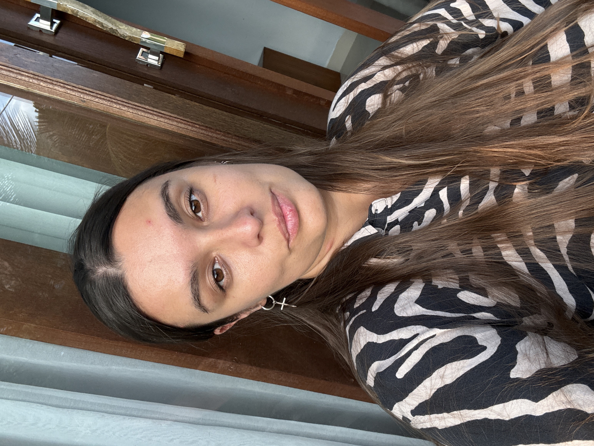 A woman is seen in a close-up selfie shot. She has fair skin, brown eyes, and long, brown hair. She is wearing a zebra-print top. Her earrings feature a circular hoop with a small cross hanging from it. The background consists of a wooden door frame and a white curtain. The location is indeterminate but is possibly in or near Pejeng, Indonesia.