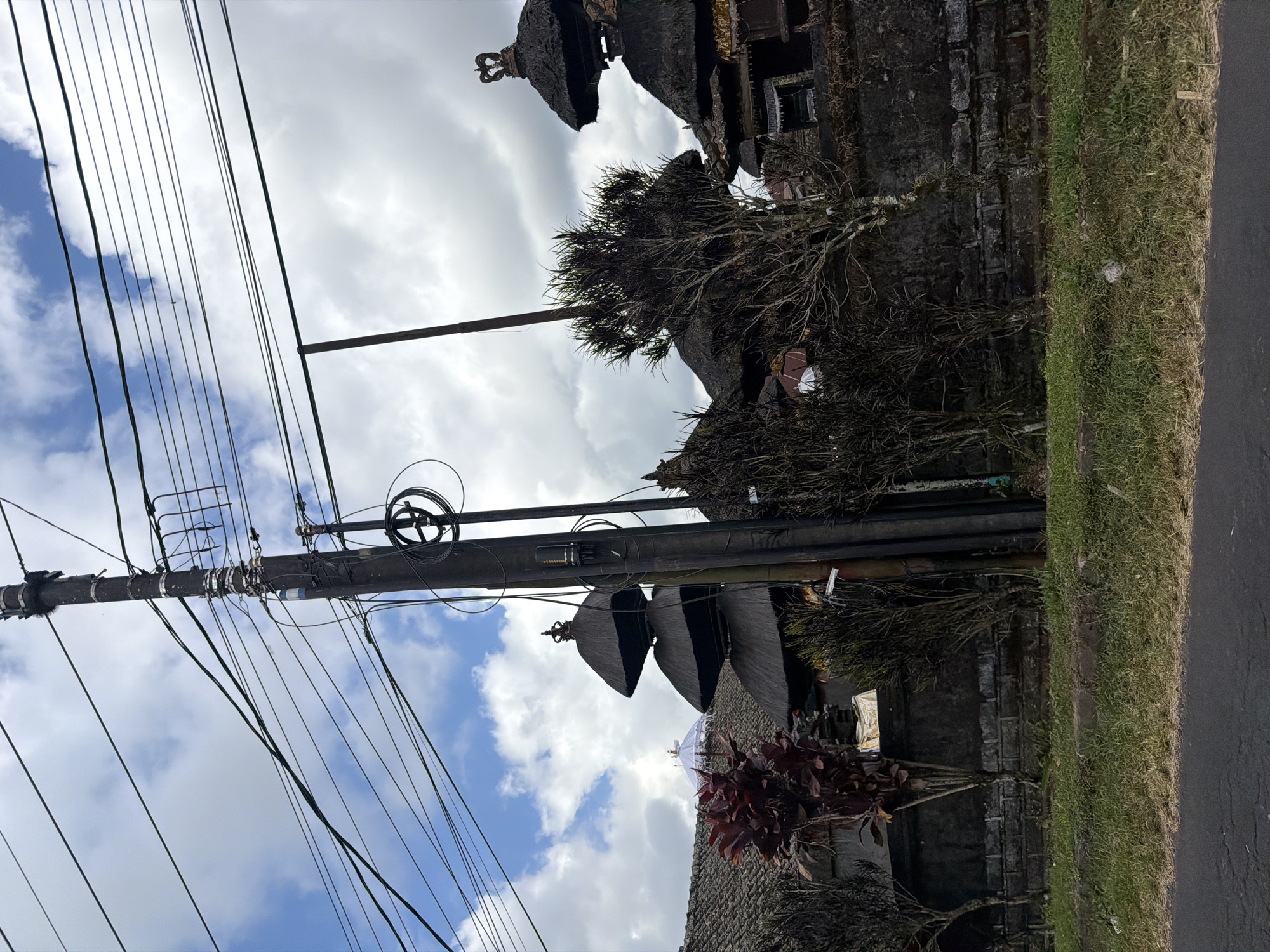 The image depicts a street view in Kintamani, Indonesia. A dark, weathered utility pole stands prominently in the foreground, adorned with numerous electrical wires crisscrossing the sky. Behind the pole, traditional Balinese architecture is visible, characterized by thatched roofs and stone walls. Specifically, tiered structures topped with ornate crowns suggest a temple or a significant cultural site. Lush green grass lines the roadside, bordering a paved road. The sky is a mix of blue and white clouds, indicating daytime. A few trees with sparse foliage are scattered throughout the scene, and a vibrant red-leafed plant adds a splash of color against the darker stone wall. The overall atmosphere suggests a tranquil, culturally rich environment.