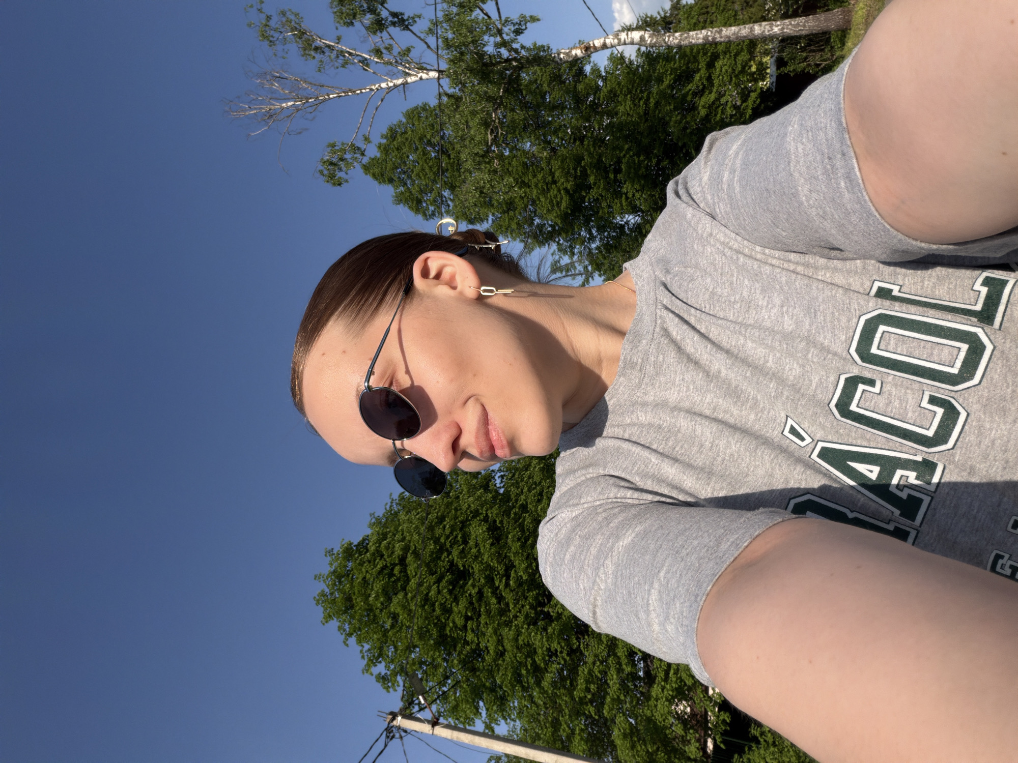 A woman is pictured from a low angle in an outdoor setting. She is wearing a gray T-shirt with green and white lettering, and round sunglasses. Her hair is pulled back. The background includes a clear blue sky, green trees, and electrical poles. The location is suggested to be Reshetnikovo, Russia.