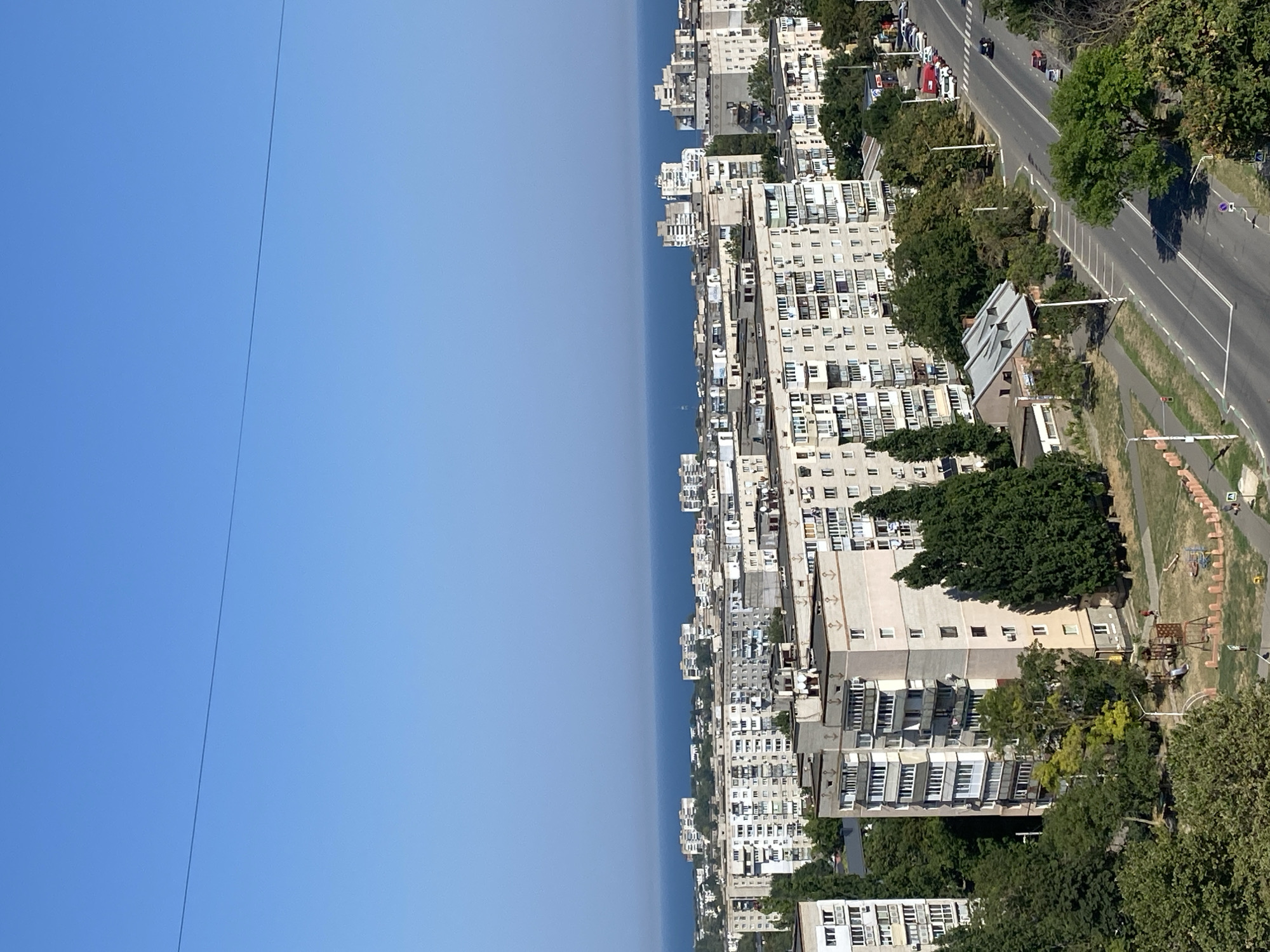 The image is a high-angle, wide shot of a cityscape in Chornomorsk, Ukraine, with the Black Sea visible in the distance. The foreground features a multi-lane road with sparse traffic, including several cars and a red truck. Adjacent to the road are sidewalks and green spaces with trees and a playground. Several apartment buildings, typical of Soviet-era architecture with light-colored facades and balconies, dominate the midground and background. The sky is a clear, bright blue with a single, thin power line stretching across the top of the frame. The overall impression is of a sunny day in a residential area of a coastal city.