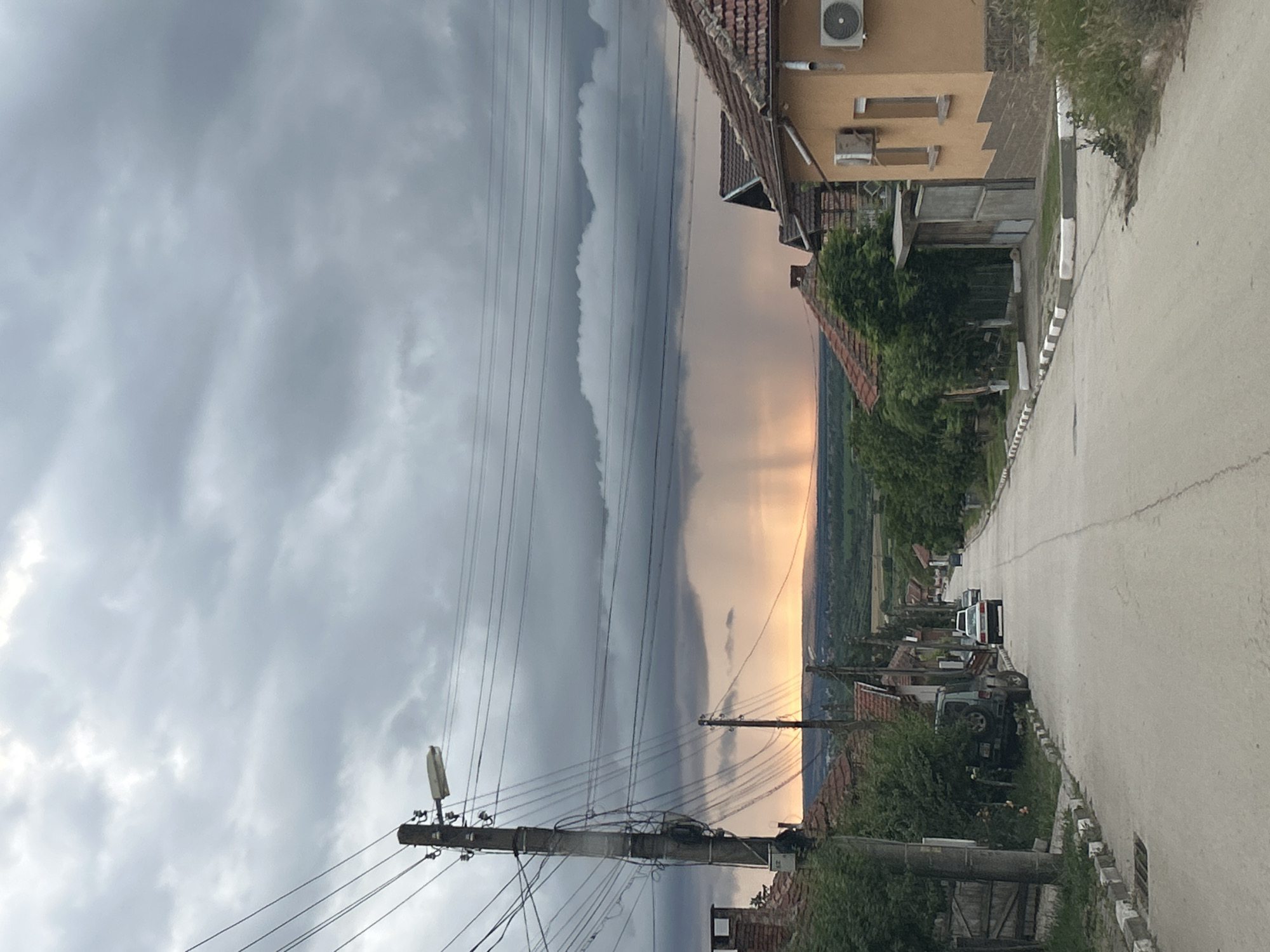 The image depicts a street scene in Milkovitsa, Bulgaria, under a dramatic, overcast sky with a visible rain shower in the distance. The perspective is from a street looking down a gentle incline. On the left, a tall utility pole with multiple wires and a streetlamp is prominent. Adjacent to the pole are houses with red tile roofs and some vegetation. A dark-colored SUV is parked on the side of the road. The street itself is paved and bordered by curbs with alternating white and black markings. Several other houses line the right side of the street, one of which is painted a light orange and features two air conditioning units on its exterior. Further down the street, two cars are visible, one white sedan and another vehicle behind it. The background shows a hazy landscape under the stormy sky, with the rain shower creating a visible curtain of water against a backdrop of warm, sunset-like light. The overall atmosphere is moody and indicative of impending or ongoing weather activity.