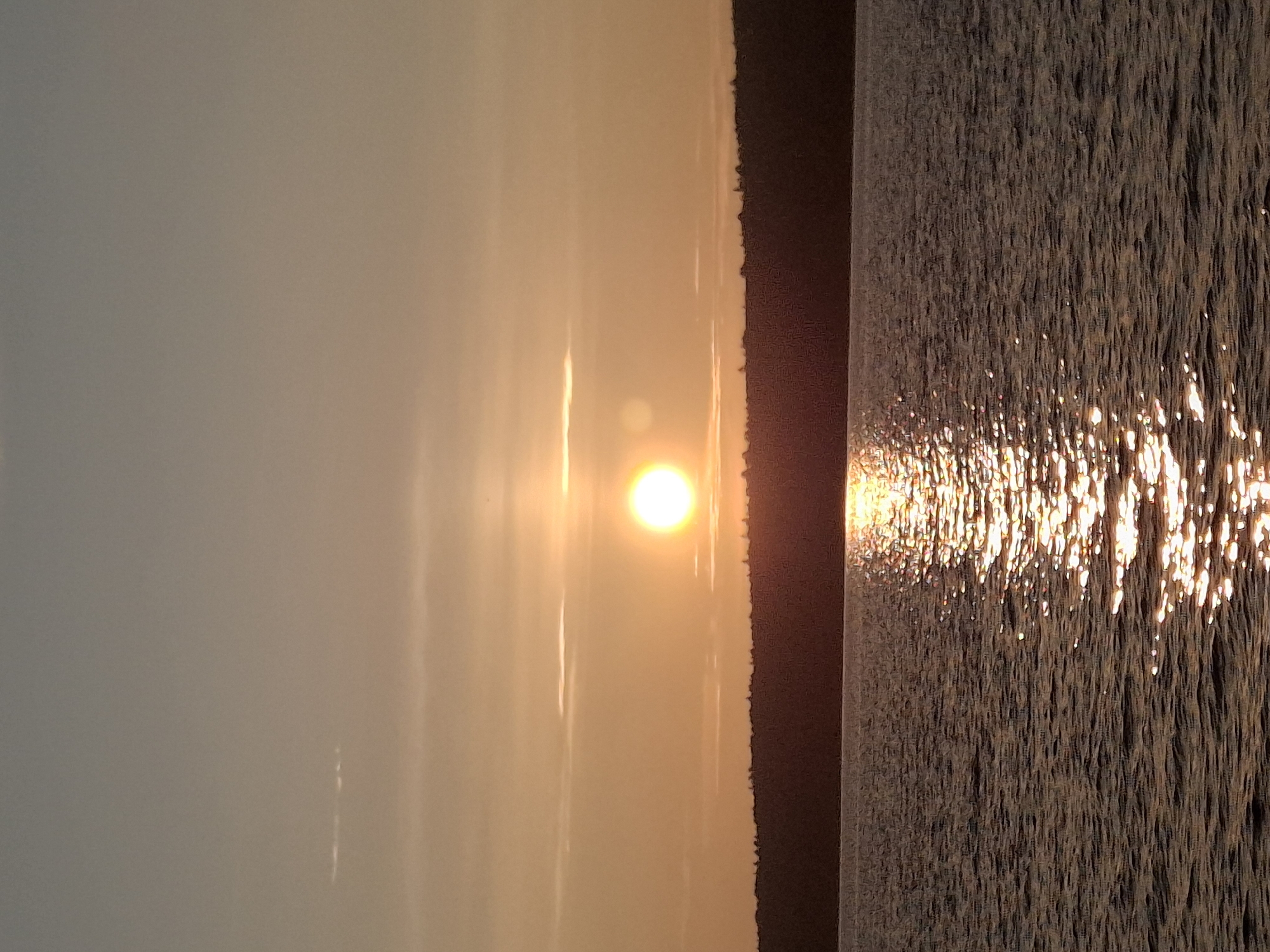 A sunset over the Kama River in Chaikovsky, Russia, illuminates the sky with warm orange and yellow hues. The sun, a bright, glowing orb, is positioned slightly left of center in the upper half of the frame, casting a shimmering reflection on the rippling water below. A dark, silhouetted treeline forms the horizon, separating the sky from the river. The water is a deep, textured blue-gray, with the sun's reflection creating a sparkling pathway of light that stretches towards the viewer. The sky is mostly clear, with a few wispy clouds scattered horizontally across the upper portion. The overall scene is one of natural beauty and tranquility.