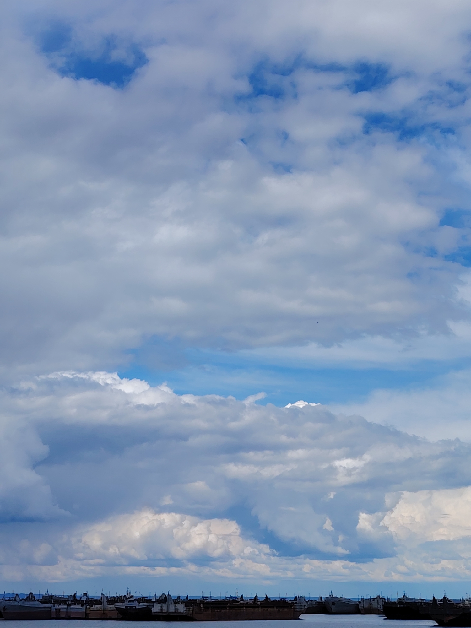 The image shows a partly cloudy sky over a body of water in Chaikovsky, Russia. In the foreground, several docked boats or ships are visible on the water. The sky is predominantly filled with white and gray clouds, with patches of blue sky visible in between. The overall impression is one of a cloudy day over a port or harbor in the city.