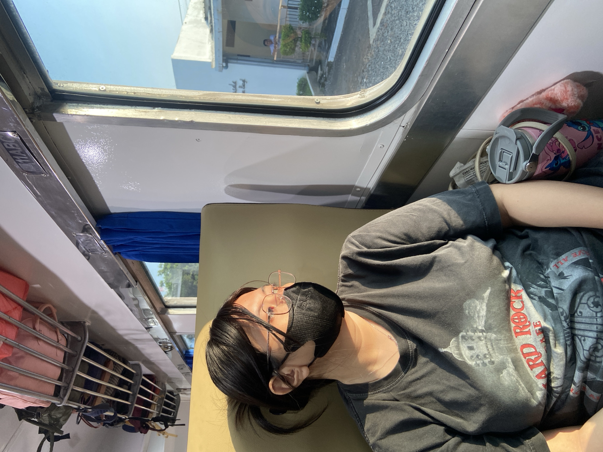 A woman is reclining on a seat inside a train. She wears glasses and a black face mask. She has dark hair and wears a dark t-shirt with red lettering that says "HARD ROCK." A water bottle with a cartoon print and a pink slipper are located next to her head. The train's window is to her left, with a building and a glimpse of the outside visible through it. A luggage rack is above her head, filled with bags. The interior of the train appears worn. Given the location, the scene is most likely in the city of Kidul Dalem, Indonesia.