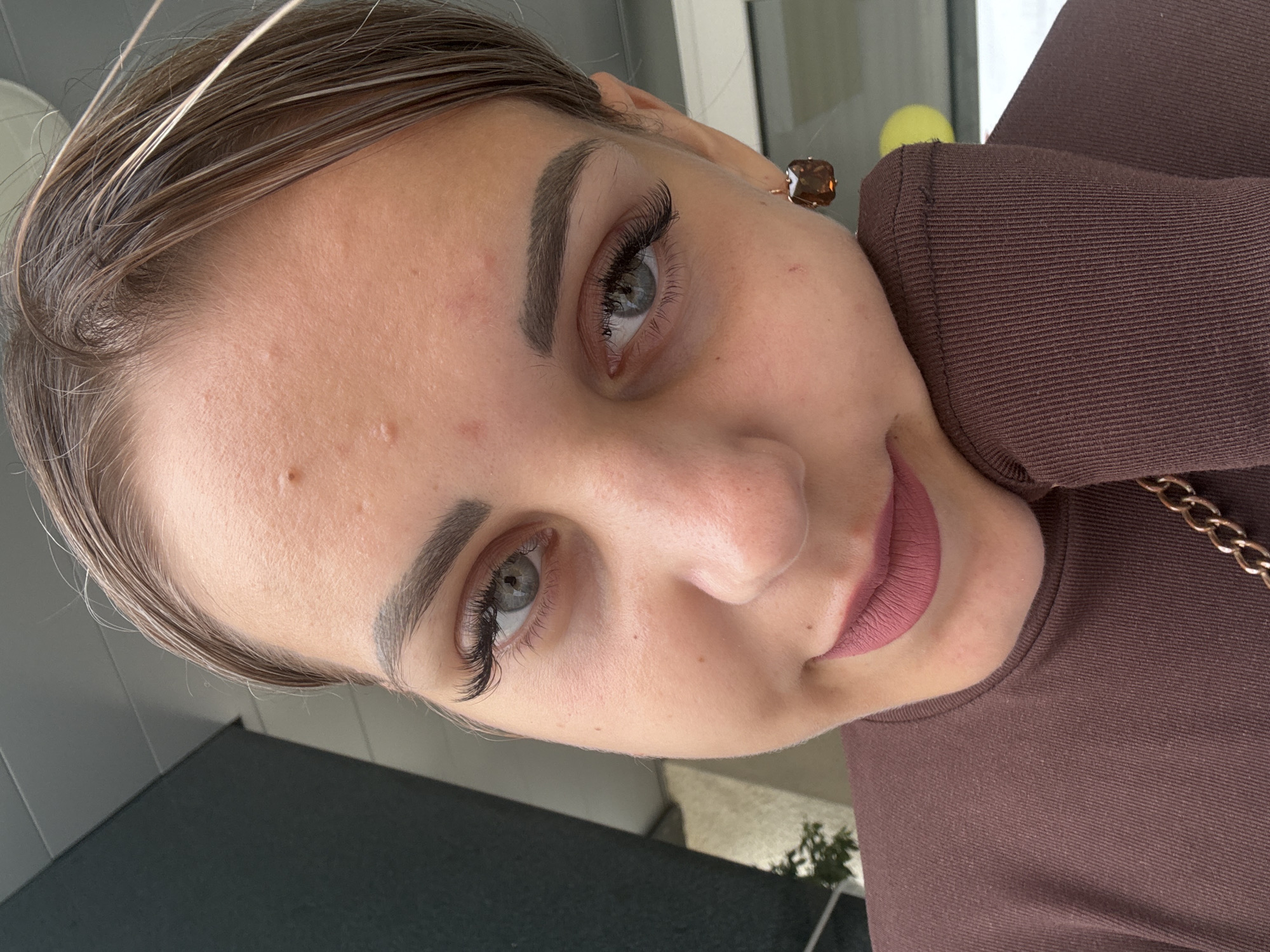 A close-up selfie shows a young woman with light brown hair, her face angled towards the camera. She is wearing a brown long-sleeved top and has dark eyeliner and mascara on her eyes, with prominent false eyelashes. Her lips are painted a matte pinkish-brown shade. She is wearing a square, brown, faceted earring in her right ear. Her left hand is supporting her cheek. The background is blurred but appears to be an outdoor setting with some architectural elements and greenery visible. There is no visible indication of the city of Yuzhno-Sakhalinsk, Russia.