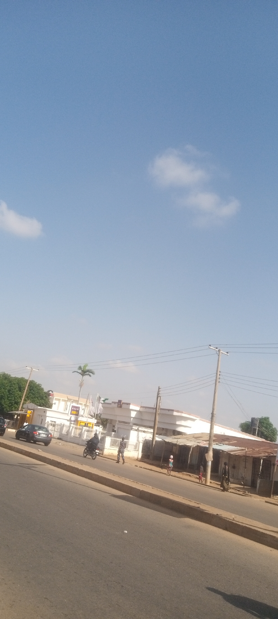 The image depicts a street scene in Sabon Tasha, Nigeria. A black sedan is parked near a white building with a sign and flags. A person on a motorbike drives along the road while an individual in camouflage walks near the building. Further down the street, several people are walking near structures with corrugated metal roofs. Utility poles and wires are visible above. The sky is light blue with scattered clouds. The road is wide with a curb separating it from the sidewalk.