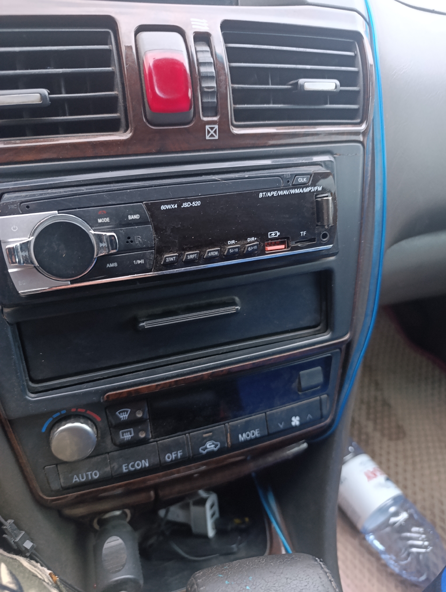 The image shows the interior of a car, specifically the center console area. A car radio is visible, with various buttons and a large dial. Below the radio are storage compartments and climate control buttons, including knobs for temperature and fan speed, as well as buttons labeled "AUTO," "ECON," and "OFF." There is also a power outlet. The overall condition of the interior appears to be used. A bottle of water is visible on the passenger side floor. The context of this image being in the city of Явлен ауылдық округі, Kazakhstan, is noted, but no specific visual information in the image directly supports or contradicts this location.