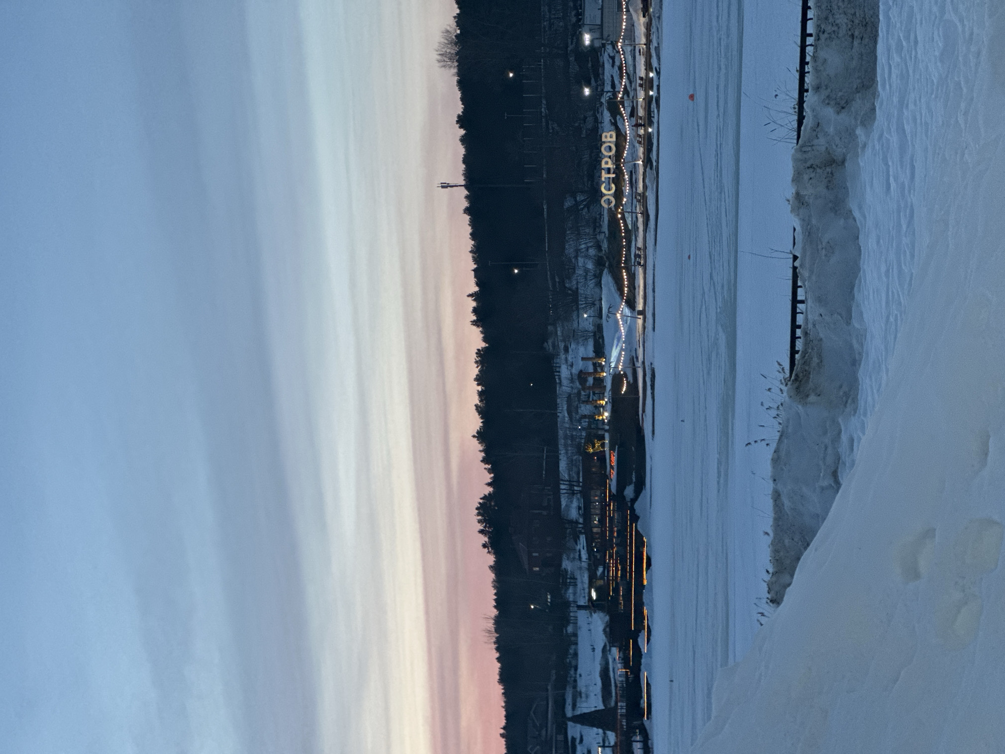 The image depicts a winter twilight scene overlooking a frozen body of water in Votkinsk, Russia, specifically focusing on the "ОСТРОВ" (OSTROV) recreational area on the Votkinsk Pond.
In the foreground, a large, uneven accumulation of snow and ice occupies the lower left quadrant, forming a bank. To the right, smaller mounds of snow and ice are present, interspersed with visible dry reeds or tall grasses protruding from the frozen ground. A dark, low-lying structure, possibly a railing or barrier, is partially obscured by snow on the immediate bank.
The midground is dominated by the frozen surface of a large water body, covered in a smooth layer of ice and snow. Two small, indistinct orange objects, potentially buoys, are visible on the ice in the distance.
The background reveals the opposite shore, featuring a developed recreational area under artificial illumination. A large, illuminated sign spelling "ОСТРОВ" (OSTROV), meaning "Island" in Russian, is prominently displayed on the snow-covered bank. Decorative string lights are strung between poles along the shoreline. To the left of the "ОСТРОВ" sign, a multi-tiered structure or building is illuminated with horizontal orange strip lights. Further left, a dark, conical structure, possibly a gazebo or architectural element, stands on the snowy bank. Several small, dark features resembling benches are scattered along the illuminated shore. Beyond this recreational strip, a snow-covered hillside rises, dotted with scattered buildings and streetlights. A dense line of dark coniferous trees forms a forest along the top of the hill. A tall, thin communication tower is visible among the trees to the right.
The sky above displays a gradient from a soft pink and orange hue near the horizon, transitioning through light blue, to a deeper blue-grey at the top. Sparse, wispy clouds are present horizontally across the sky, consistent with dusk or early night conditions. No individuals are clearly visible in the scene.