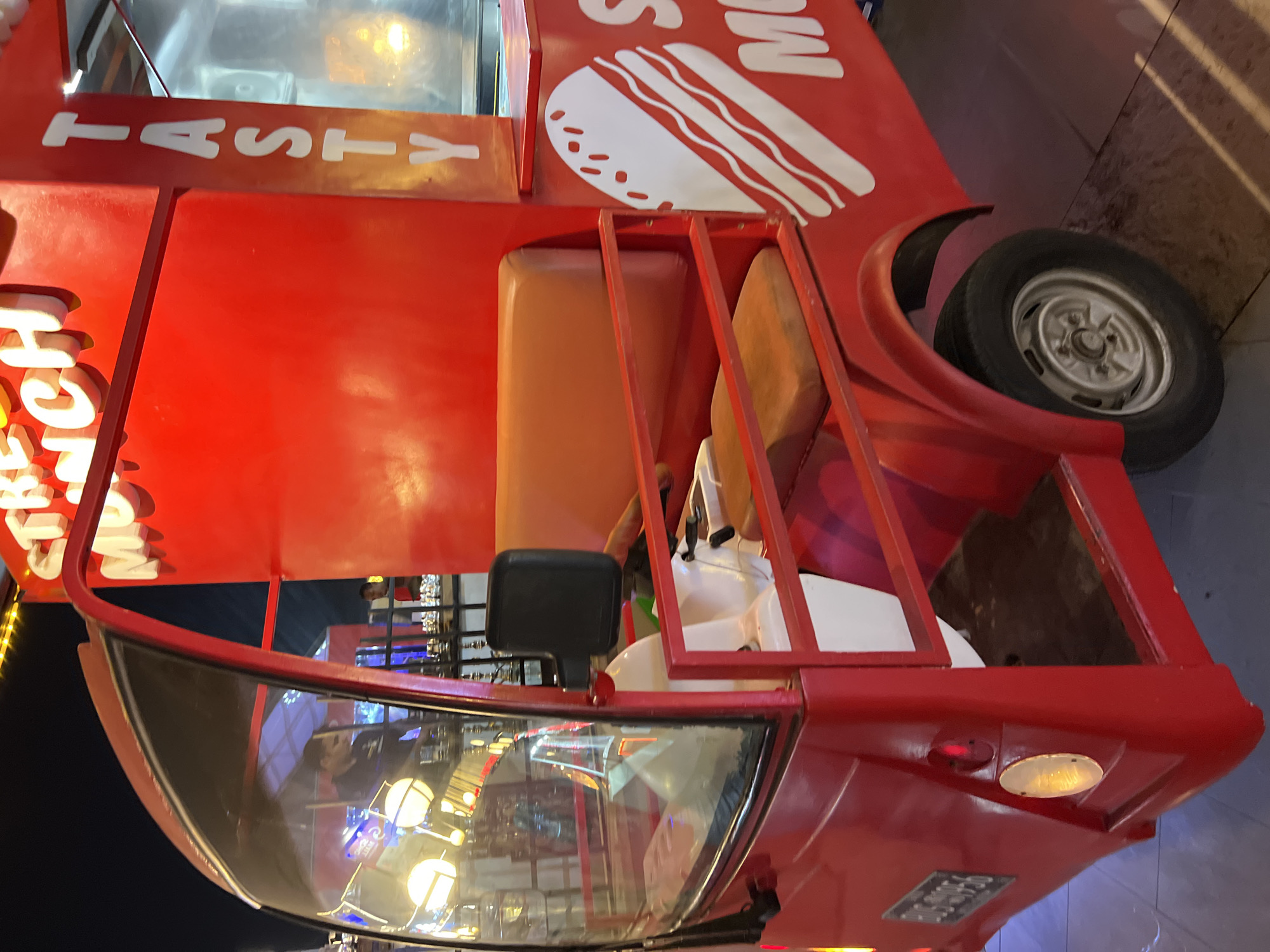 A red food truck with "STREET MUNCH" written in large white letters on top is parked at night on a tiled surface, likely in Sharm El Sheikh, Egypt. The truck has a driver's cabin with a glass windshield and a seat inside. The side of the truck features a large white graphic of a hamburger and the word "TASTY" written vertically. A person is visible inside the driver's cabin, and another person appears to be working behind the counter at the rear of the truck. The background shows a dark night sky with illuminated string lights and what appears to be other structures or stalls. The truck has wheels and a license plate visible on the rear fender.
