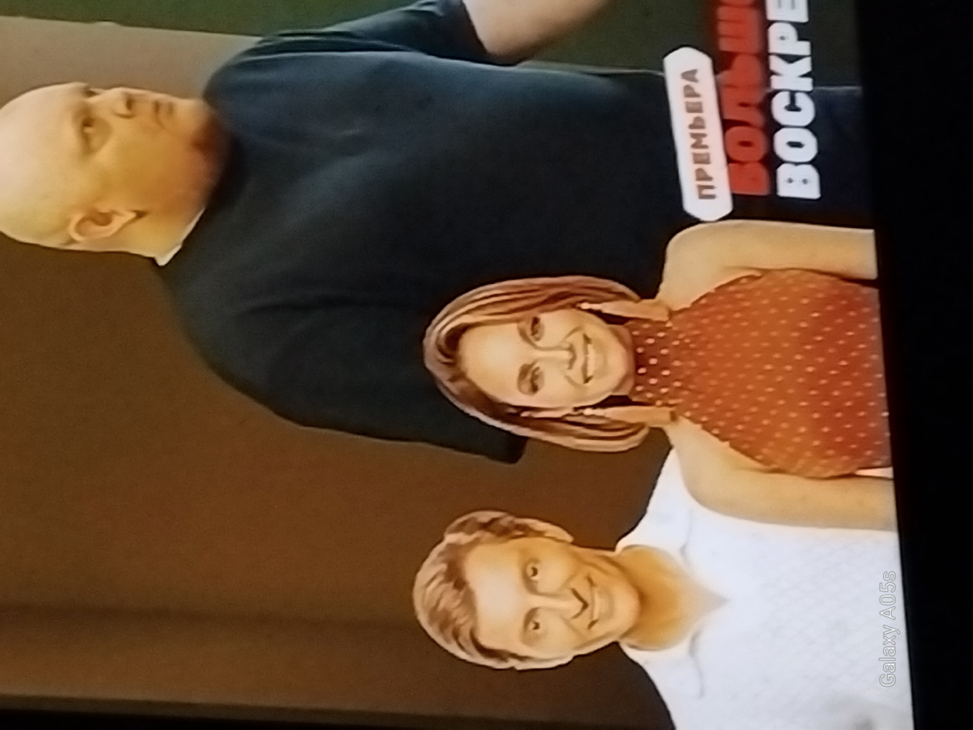 The image displays a posed photograph of three individuals. On the left is a light-skinned male with short, styled brown hair, a white collared shirt, and a wide smile. Next to him, in the center, is a light-skinned female with shoulder-length blonde hair, wearing a red polka-dot halter top and large earrings. She is also smiling. Behind them and to the right is a bald, light-skinned male with a thick build, wearing a dark crew-neck shirt. In the background, a dark brown wall is visible. Partially obscured text on the right reads "ПРЕМЬЕРА" and "ВОСКР". A watermark in the bottom left corner indicates "Galaxy A05s". There is no indication of the city of Druzhba, Russia, or any specific urban environment in this image.