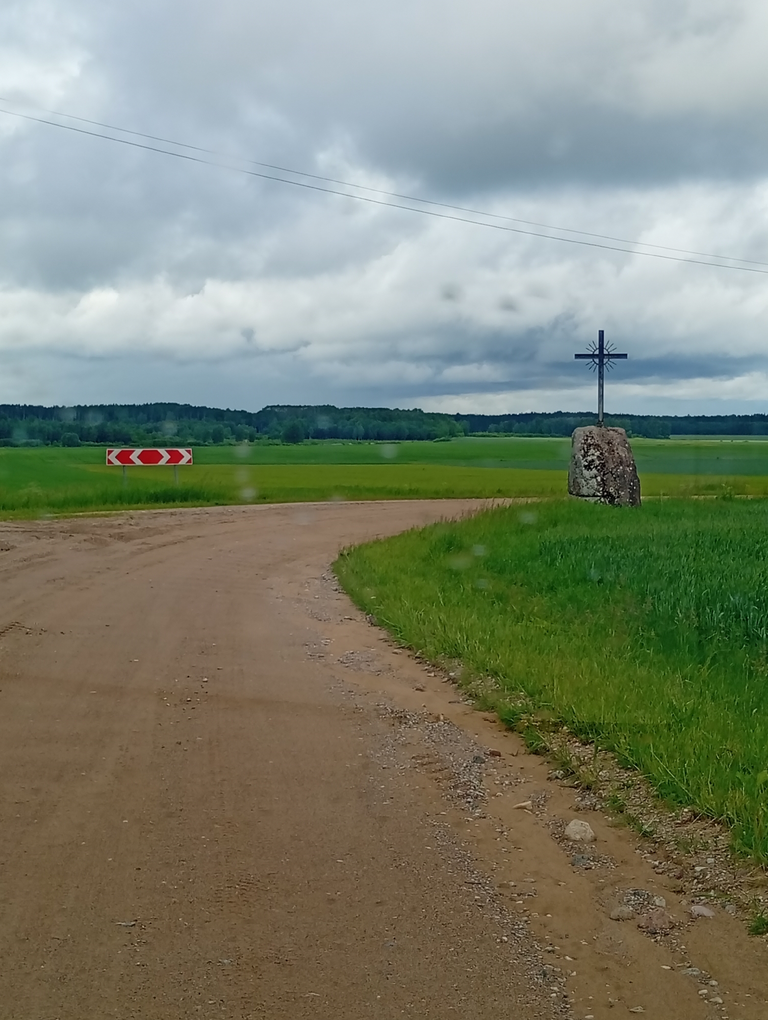 The image depicts a rural landscape in Krikliai, Lithuania, under an overcast sky. A dirt road curves to the left, leading towards a field of green grass. On the right side of the road, a large rock stands with a metal cross on top, its silhouette sharp against the cloudy sky. A traffic sign with red and white chevrons indicating a sharp turn is visible to the left, in front of the green field. The scene appears to be an outdoor, natural environment.