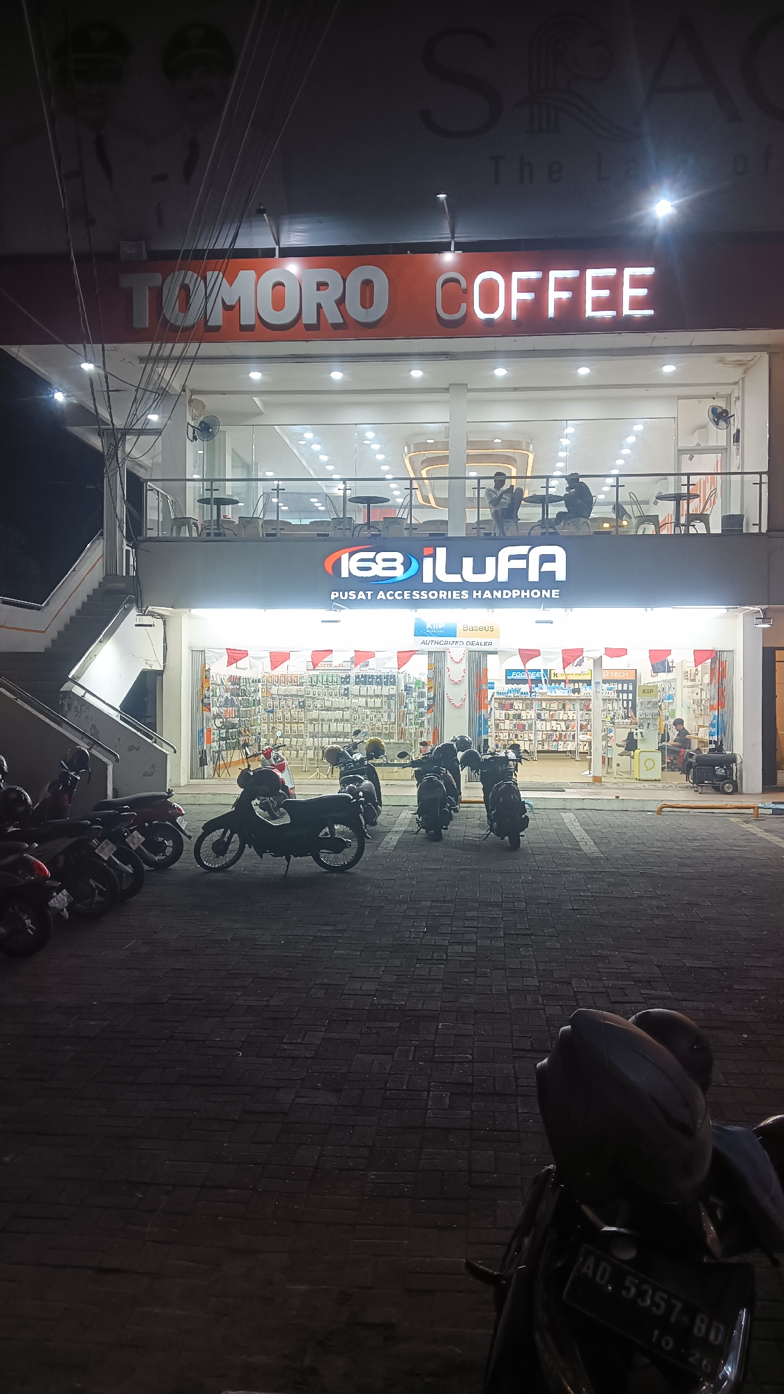 The image shows a night time exterior view of a commercial building in Sragen, Indonesia. The building features a prominent orange sign with white lettering that reads "TOMORO COFFEE". Below this sign, another sign displays "168 iLUFA" and "PUSAT AKSESORIS HANDPHONE", indicating a mobile phone accessory store. The storefront is illuminated, revealing shelves stocked with merchandise. Several motorcycles are parked on the paved area in front of the building. In the upper part of the building, there is a balcony area with tables and chairs, where a few people are seated. The overall environment suggests an urban commercial district.