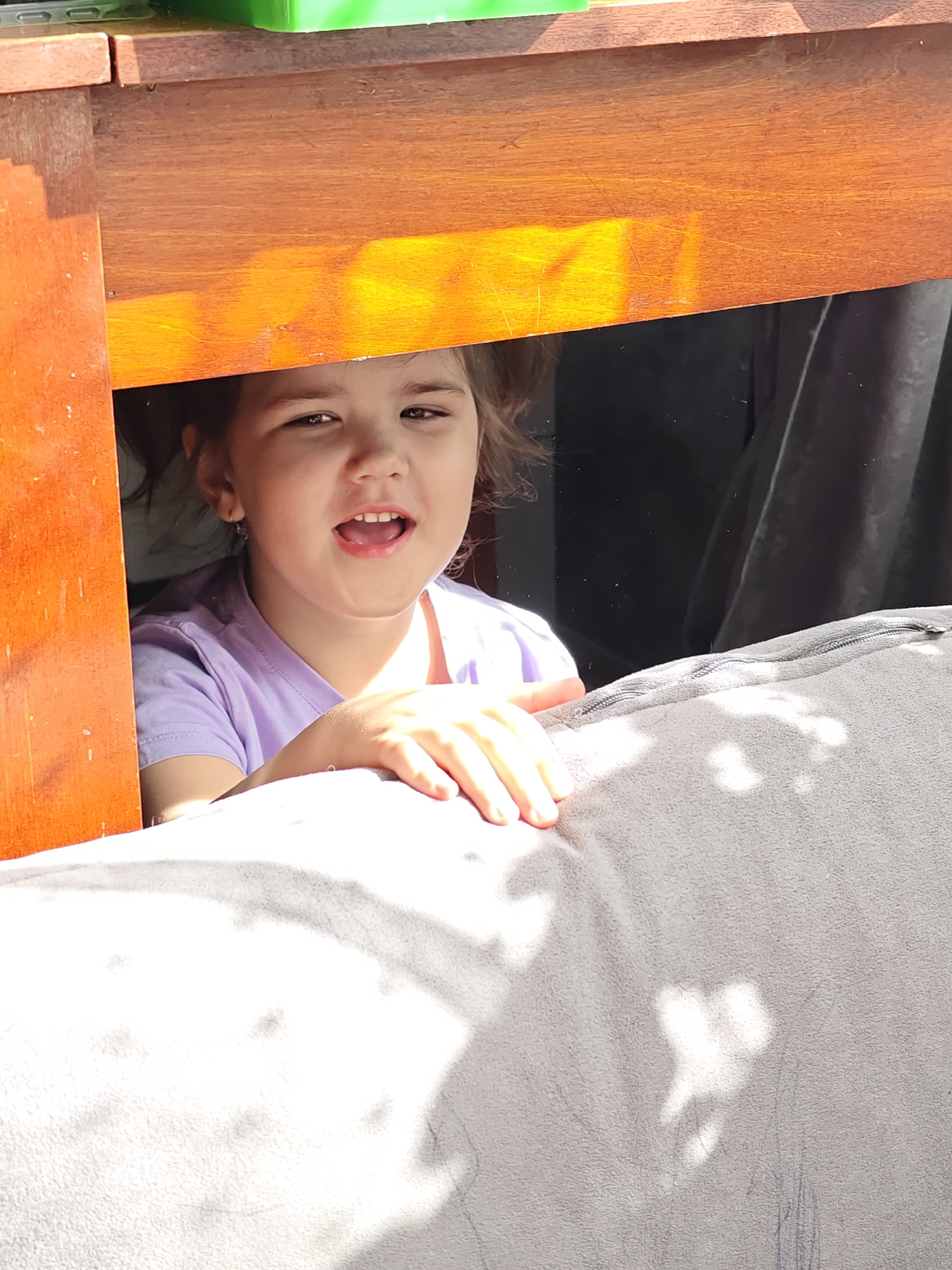 A girl with dark hair and fair skin, wearing a light purple t-shirt, looks out from under a wooden structure. Her mouth is open slightly, and she rests her hand on a gray, textured cushion or blanket. The backdrop includes dark fabric or shadows. The location is Уральское, Russia.