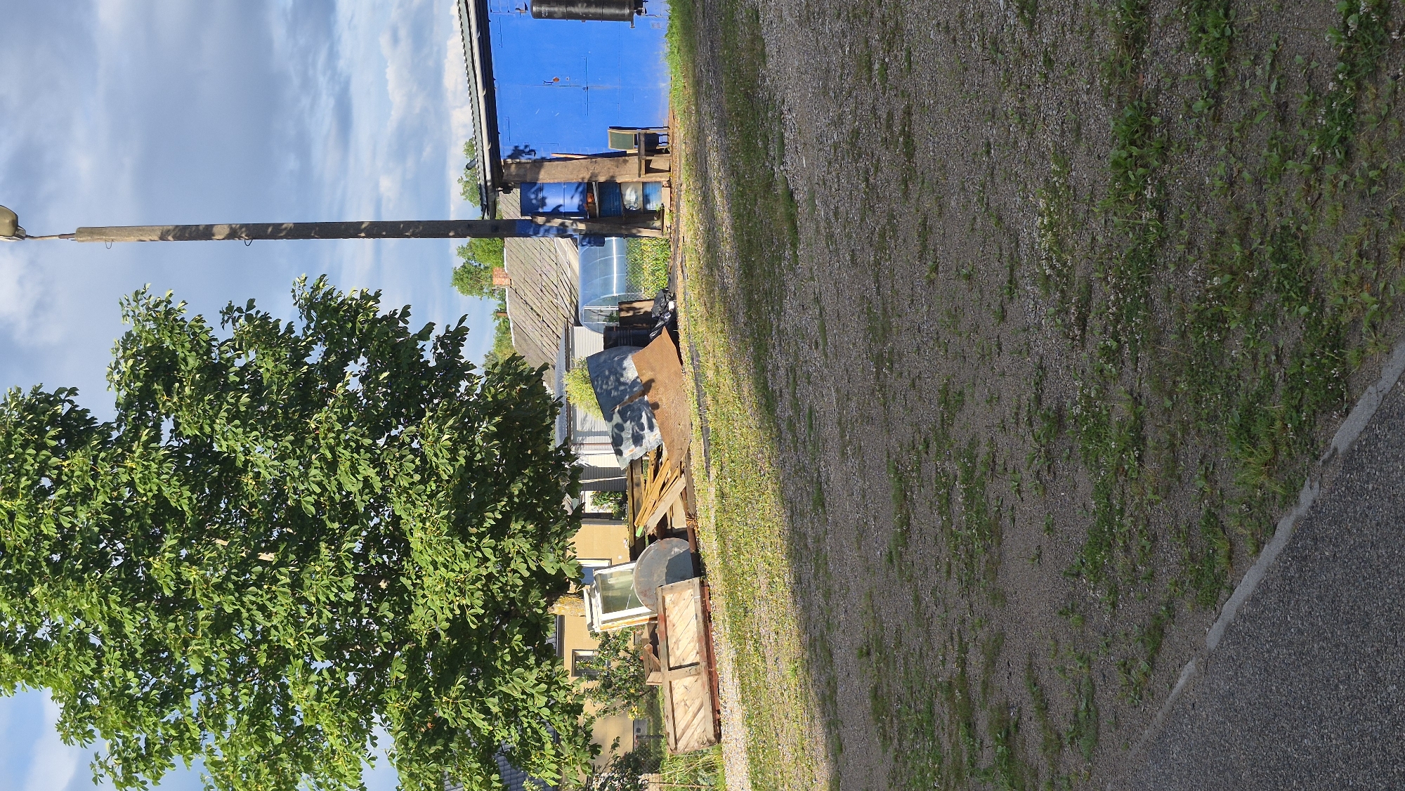 A full shot captures a scene outside, featuring a large green tree on the left, a tall wooden pole with a light fixture on top, and a bright blue metal building on the right. A pile of assorted scrap metal and wooden objects lies in the foreground. In the background, houses with grey roofs are visible, along with a small greenhouse. The ground is covered in gravel and patches of grass. The sky above is partly cloudy.