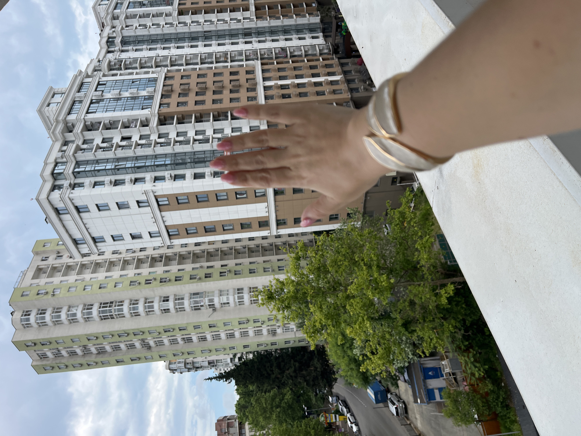 In Sochi, Russia, a first-person perspective captures a view of residential buildings and a street. A woman's hand, adorned with pink nail polish and a bracelet, is extended towards the camera, obscuring part of the scene. Behind her hand, a tall, slender, pale green building with numerous balconies and windows dominates the left side of the frame. To its right, a modern, multi-story building with a white facade, brown accents, and large, reflective windows stands out. Below these buildings, a street is visible with several parked cars, including a silver sedan and a blue trailer. Lush green trees and foliage fill the midground, softening the urban landscape. The sky above is overcast with scattered clouds.