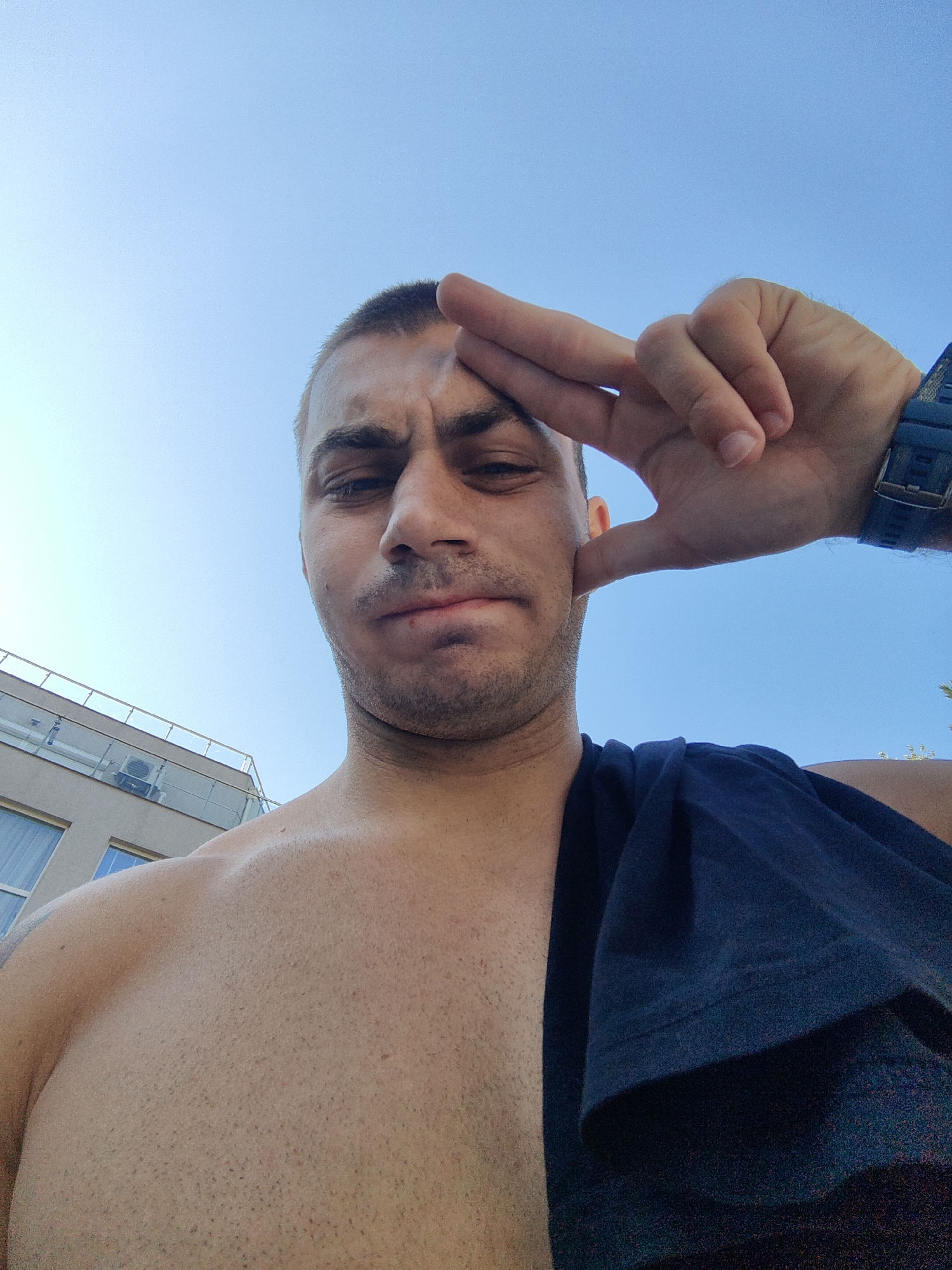 An outdoor, low-angle, selfie-style photograph captures a shirtless Caucasian male, appearing to be in his late 20s or early 30s, against a clear, pale blue sky. He has short, dark hair and a light stubble. His left hand is raised to his forehead, with his index finger and thumb touching his temple, while his other fingers are extended upwards, forming a V-shape. He wears a dark blue watch on his left wrist. A dark blue t-shirt is draped over his right shoulder. His facial expression is difficult to discern clearly due to the lighting and angle. In the background, a light-colored building with balconies and an air conditioning unit is visible to the left, suggesting an urban or resort setting. This is likely an exterior shot in the city of Varna, Bulgaria, given the architectural style and common environments.