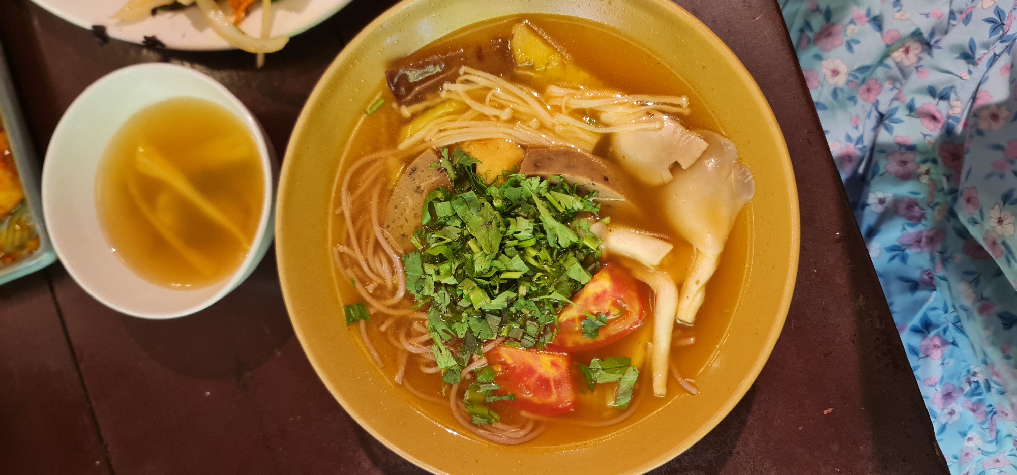 A high-angle shot depicts a bowl of Vietnamese noodle soup, possibly Mi Quang, set on a dark wooden table next to a small bowl of broth. The soup contains rice noodles, various types of mushrooms (including enoki), slices of Vietnamese sausage, tomato wedges, and is garnished with chopped cilantro. The soup is in a light brown broth. The soup is served in an earthenware bowl. A floral-patterned cloth sits beside the table. The location is likely in Đà Nẵng, Vietnam.