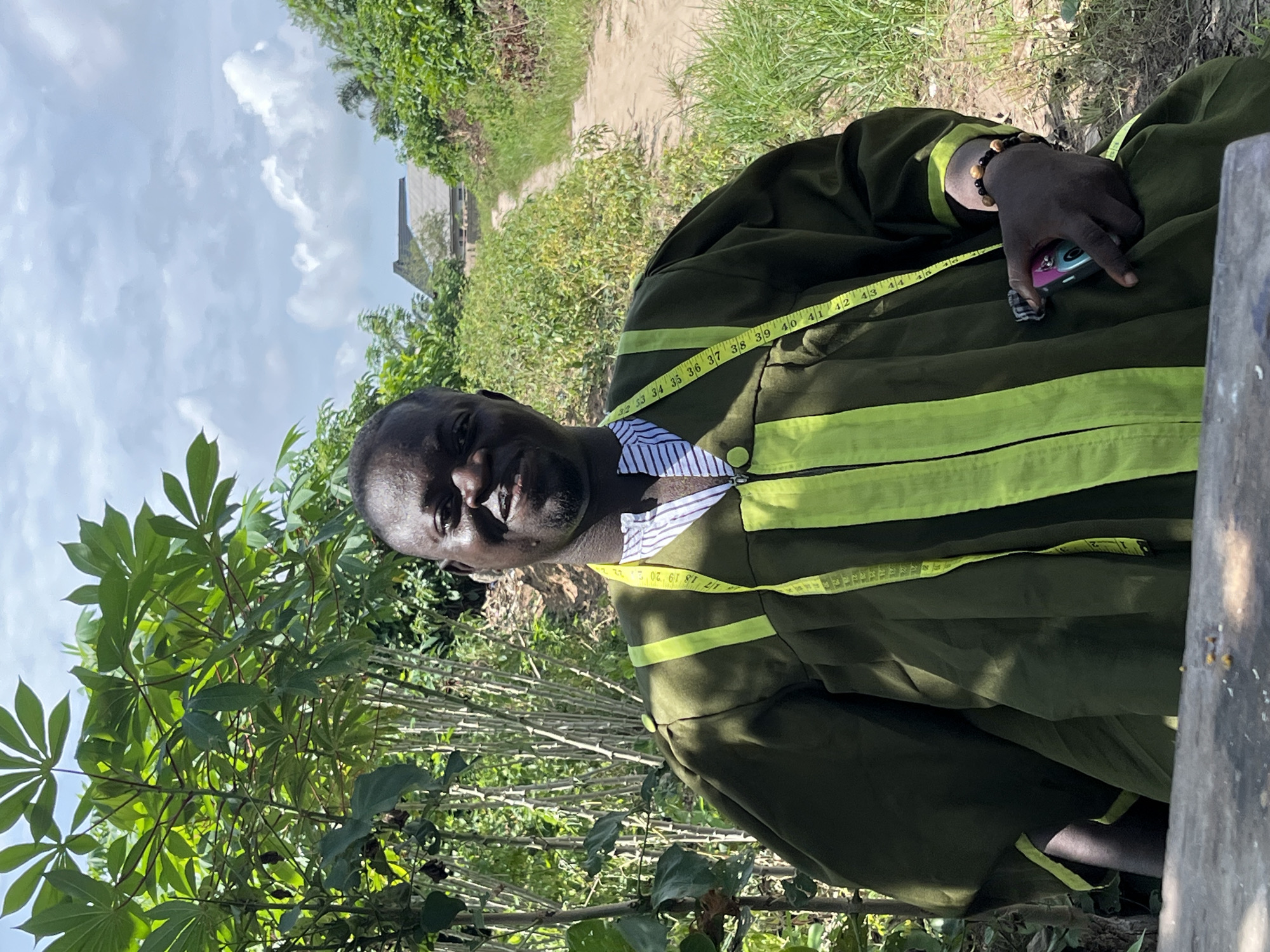 An eye-level, angled portrait photograph shows an adult Black man with a tape measure slung around his neck, apparently in an outdoor area in the city of Tofa, Nigeria. He is dressed in a garment with green and chartreuse stripes. The subject is seated, holding a small item in his hand. In the background are a dirt path, low-lying vegetation, and a cluster of trees. The sky is visible in the upper portion of the frame.