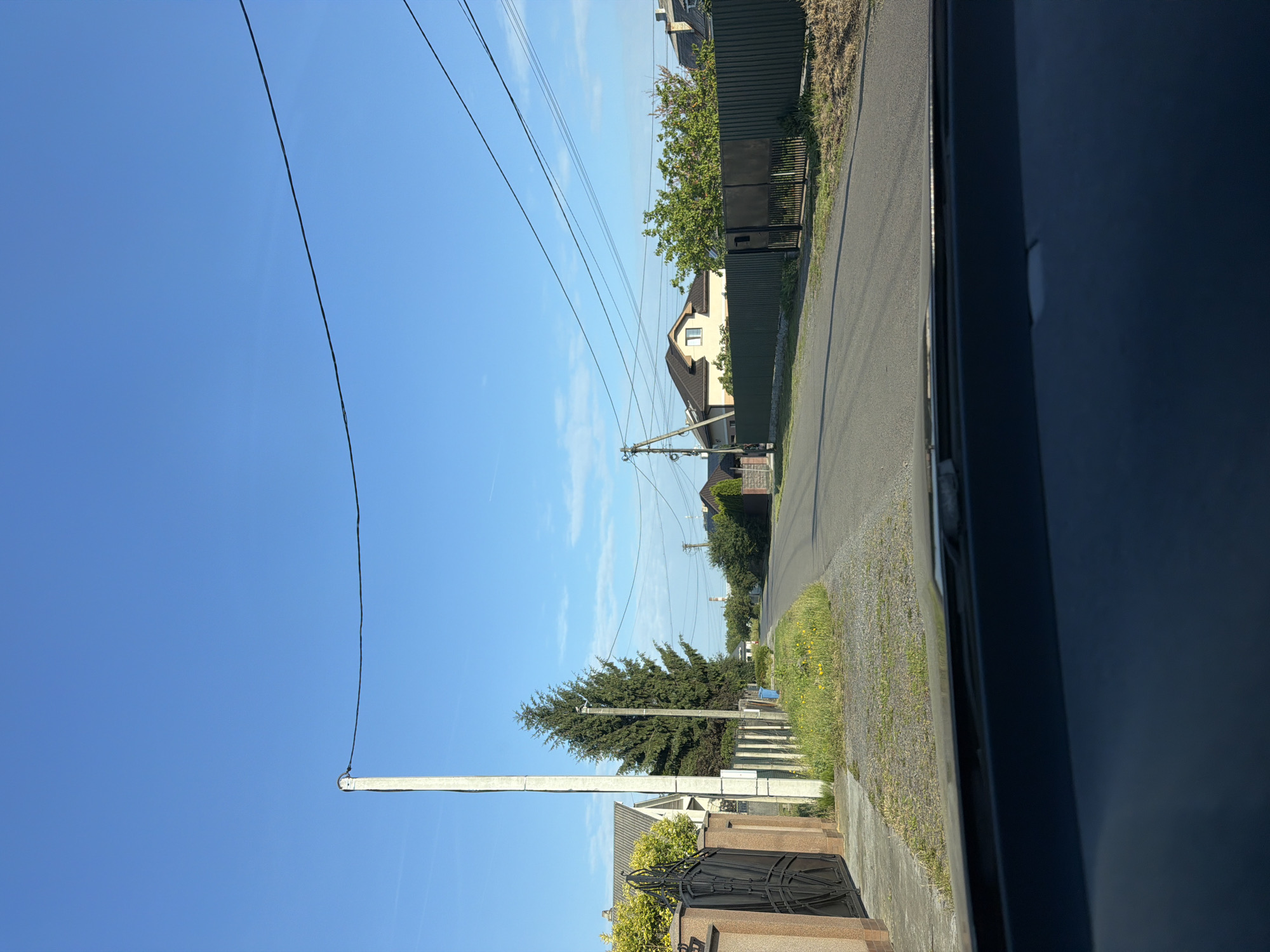 The image shows a paved road in the city of Rozivka, Ukraine, on a clear day with a bright blue sky. The road is lined with houses and fences. On the left side of the road, there is a tall concrete utility pole with power lines running across the sky. Beside the pole is a black metal gate and a beige-colored fence. A large evergreen tree stands behind the fence. On the right side of the road, there are more houses, a green metal fence, and trees. The road is empty of vehicles and pedestrians. The scene appears to be a residential street.
