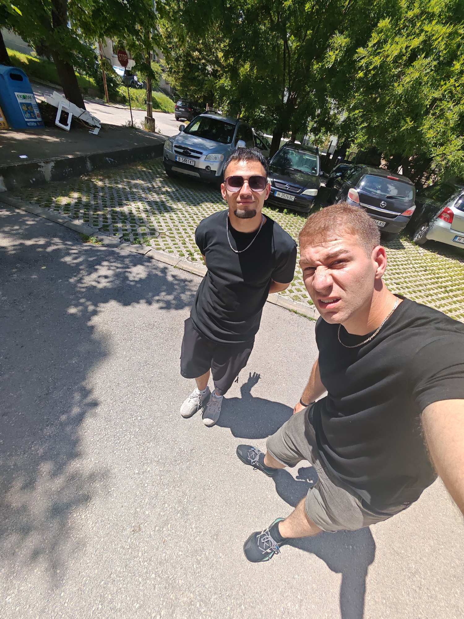 Two young men are standing on a paved street in Shumen, Bulgaria, under a canopy of trees. The man on the left is wearing sunglasses, a black t-shirt, and grey sneakers. He has a goatee and is wearing a silver chain. The man on the right is looking directly at the camera with a grimace on his face. He is wearing a black t-shirt and grey shorts, and black sneakers with blue laces. He is also wearing a silver chain. In the background, there are several parked cars and a concrete parking lot. The scene is brightly lit by sunlight, casting dappled shadows on the ground. The area appears to be a public street with trees and a parking lot.