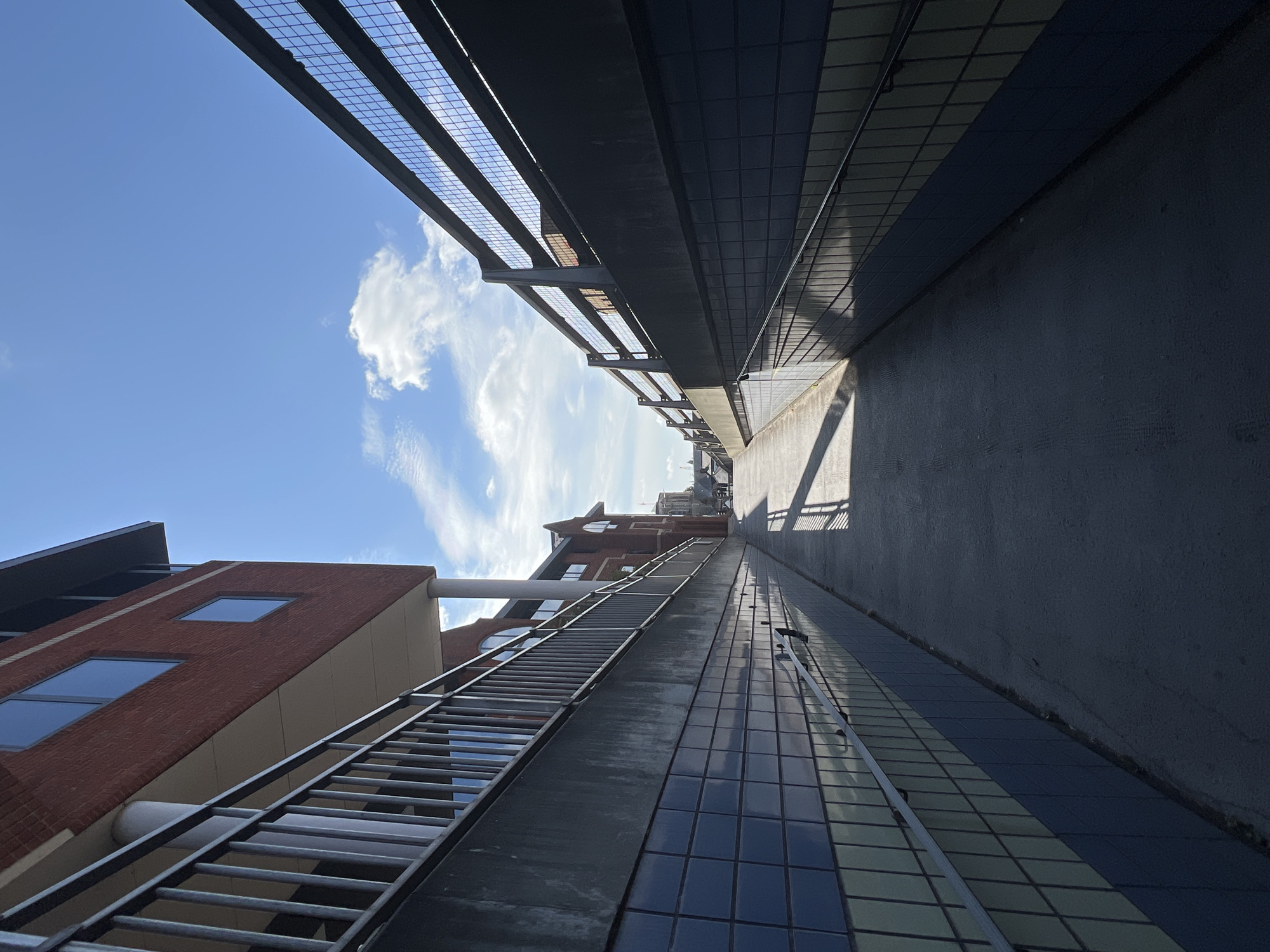 The image shows a ramp leading upwards between two walls with railings. The wall on the left is made of red brick with windows on an upper level. The lower part of this wall and the opposite wall are clad in blue and yellow tiles. The ramp itself is concrete, with a shadow cast across it from a railing structure. The sky is blue with scattered white clouds. The scene appears to be in an urban environment, possibly a pedestrian walkway or access route in Guildford, United Kingdom, given the architectural style and the presence of similar structures in the city. No people or specific actions are visible.