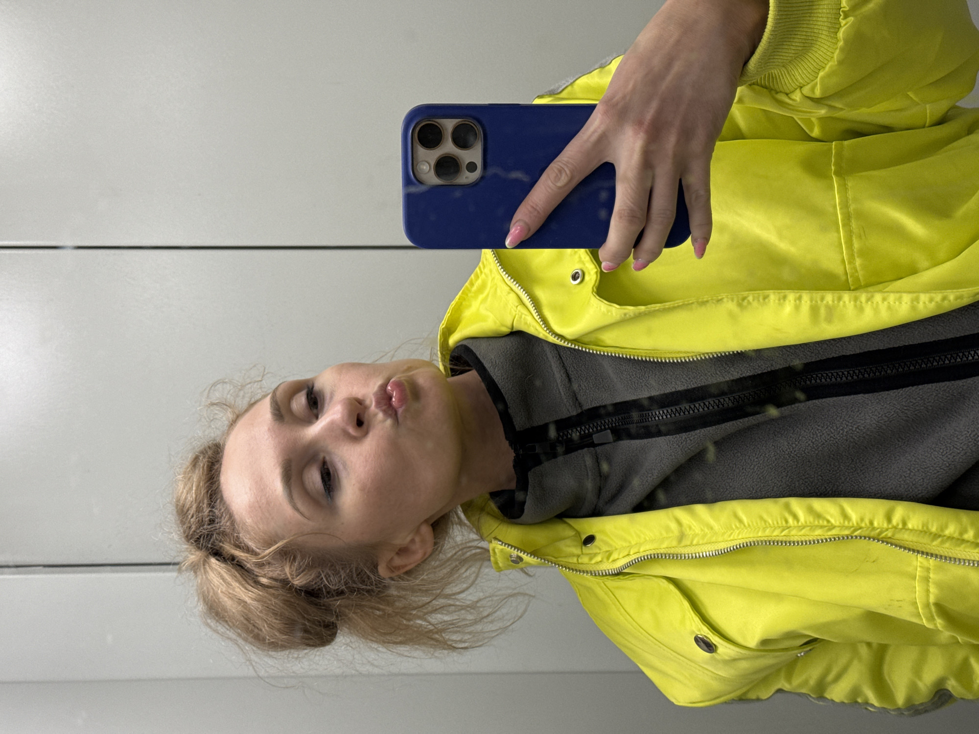A young woman is depicted in a mirror selfie, holding a blue smartphone with three distinct camera lenses visible on its back. The image is oriented sideways, with the woman's head tilted to the right. She has light brown or blonde hair, styled in a messy bun or ponytail, and is making a playful 'duck face' expression with puckered lips. Her eyes are adorned with dark eyeliner or mascara.
She is wearing a bright, neon yellow puffer jacket or windbreaker, which features a silver zipper and snap buttons. Beneath the yellow jacket, a grey fleece top with a black zipper is visible. Her left hand, holding the phone, has fingernails painted with pink polish.
The background appears to be a plain, light grey or white reflective surface, likely a mirror, with a dark horizontal line bisecting it, suggesting a seam or panel division. The overall setting is simple and uncluttered, indicating an indoor environment. No specific time of day or weather is discernible from the image. The location context provided for this image is Perm, Russia. There is no visible text in the media file.