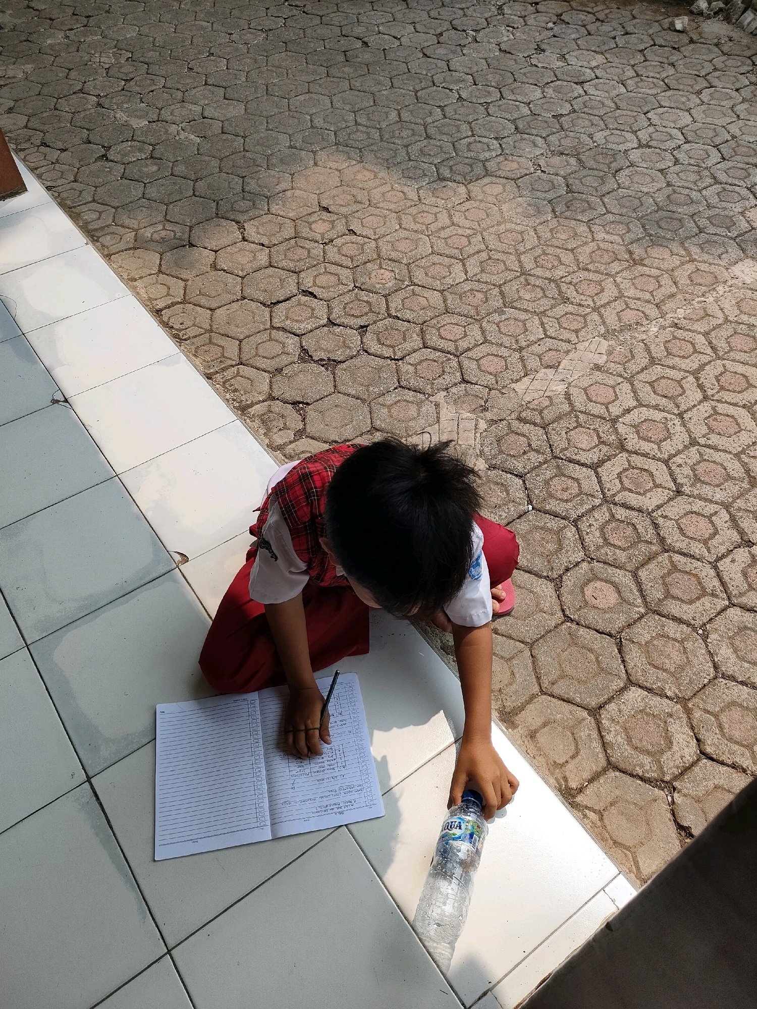 A young child, dressed in a red and white checkered school uniform with a red skirt, is sitting on tiled flooring outside. The child is facing downwards, engaged with an open notebook and a pencil, appearing to be studying or completing homework. Next to the child is a partially full plastic water bottle with the "AQUA" logo visible. The background consists of hexagonal patterned paving stones, typical of outdoor walkways or courtyards, with dappled sunlight and shadows indicating an outdoor setting. The scene is likely located in Bojong, Indonesia, based on the presence of the specific "AQUA" brand of bottled water, which is prevalent in Indonesia.