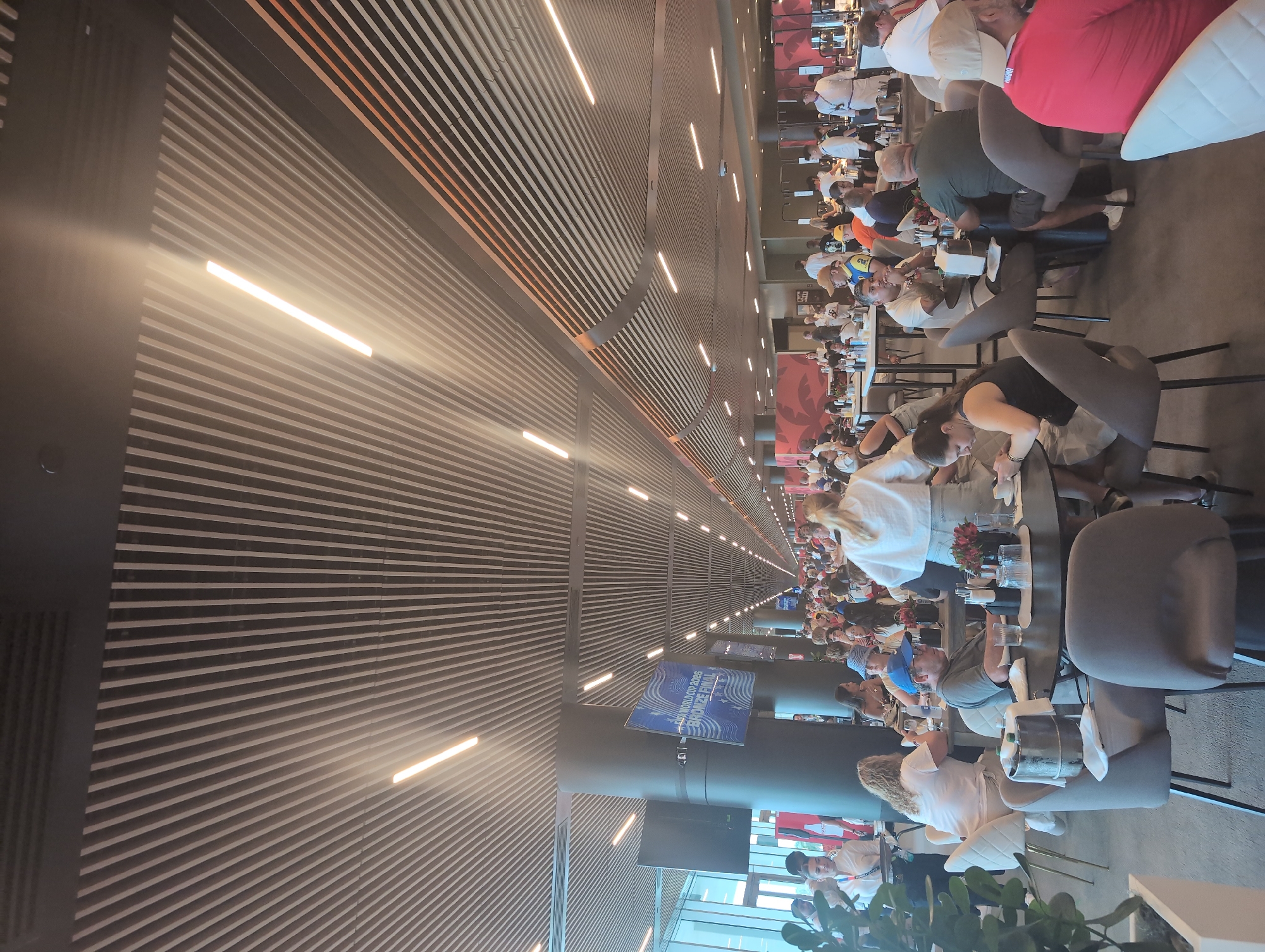 The image depicts an indoor dining area, likely within a stadium or large event venue, in Miami Gardens, Florida, USA. A long, spacious room is filled with numerous tables and chairs, many occupied by patrons. The ceiling features a modern design with slatted wood panels and integrated lighting. In the background, a large screen displays "FIFA WORLD CUP 2026 BRONZE FINAL." People are seen seated at tables, eating, drinking, and conversing. Some staff members, including individuals in white uniforms possibly resembling chefs, are visible. The overall atmosphere suggests a busy and social gathering.