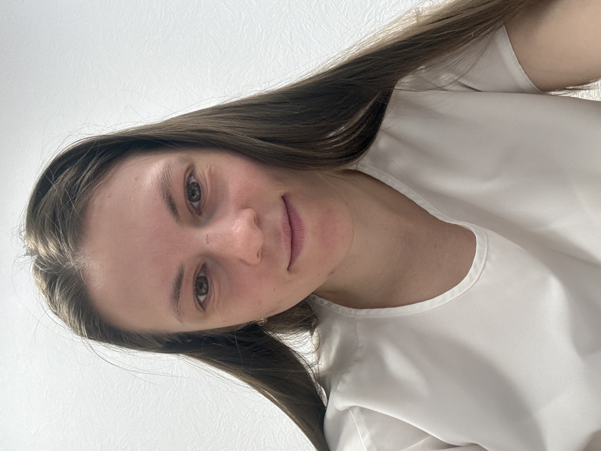 A woman with long, brown hair is shown in a close-up, tilted shot. She is wearing a white top. The background is a plain, light-colored wall. No location information is discernible in the image.