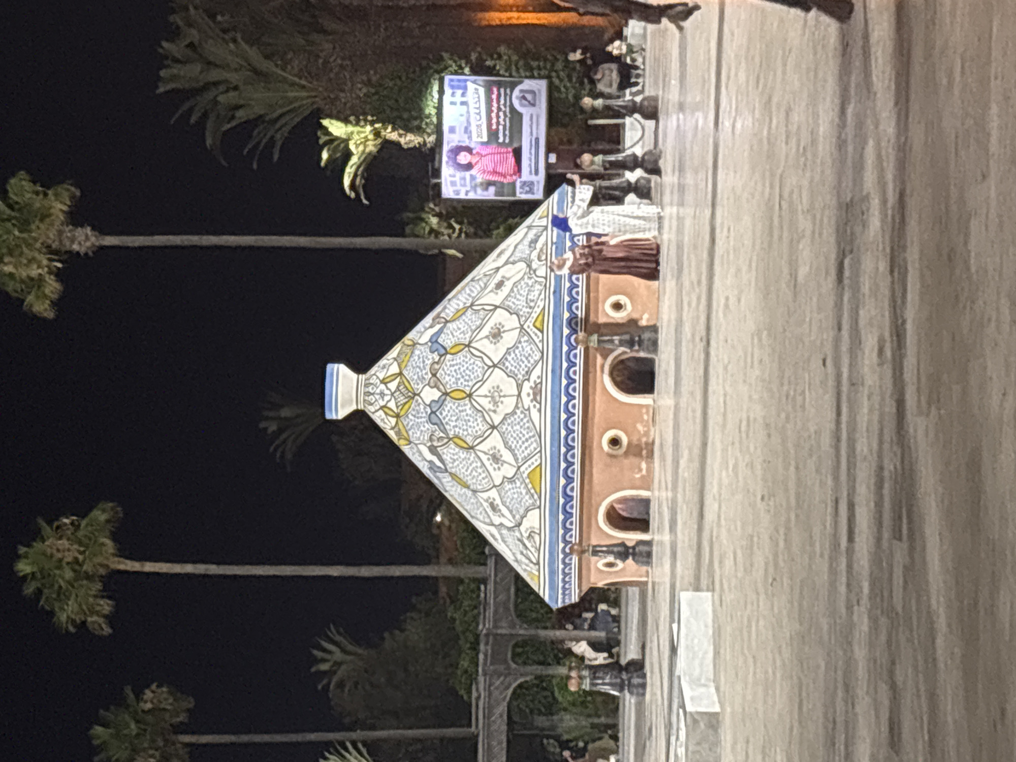 In an outdoor plaza at night, a domed structure with intricate geometric patterns and a bright blue rim is the central focus. The structure resembles a tagine, a traditional North African cooking vessel, and has arched openings at its base. Two women stand in front of it, one in a dark red dress and the other in a white patterned dress and blue headscarf, seemingly posing or interacting with a phone. To the right of the structure, a large screen displays an image of a young woman with curly hair wearing a striped sweater, with Arabic text above and below her. Tall palm trees are visible in the background, and the ground is paved. Other figures are present in the background, some seated, and more palm trees are illuminated by lights. The scene suggests an event or performance taking place in a public space.