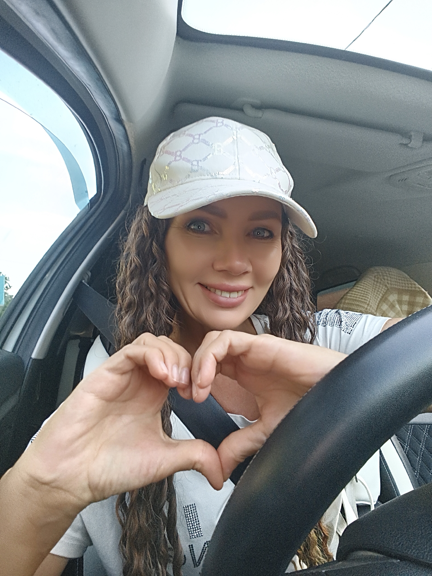 A medium shot shows a woman with long, curly brown hair and blue eyes. She is wearing a white baseball cap with a holographic pattern and a white t-shirt. The woman is inside a car, with her hands forming a heart shape in front of her chest. The steering wheel of the car is visible in the foreground. The background shows the interior of the car, including a seatbelt and part of a blanket or jacket. The woman is smiling and looking at the camera.