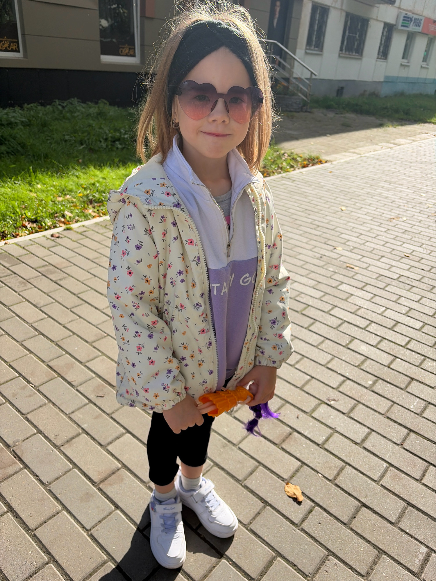 A young girl with blonde hair and heart-shaped sunglasses stands on a paved sidewalk in a city. She is wearing a floral jacket over a purple and white sweatshirt, black leggings, and white sneakers. She is holding an orange doll with purple hair. The background shows a building with windows and a grassy area with trees. The scene appears to be in the city of Chaikovsky, Russia.