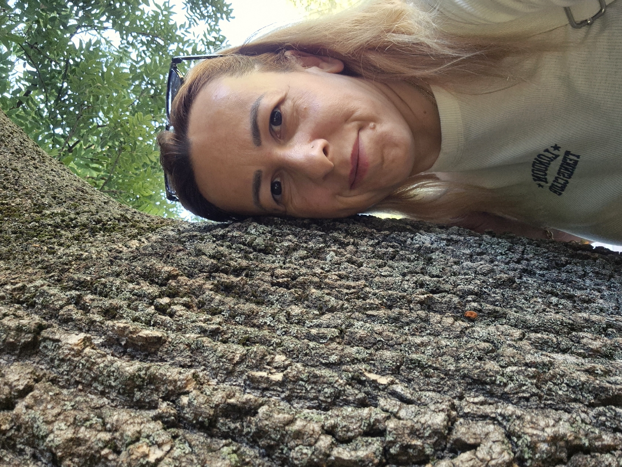 A light-skinned woman is leaning against a large tree trunk, looking at the camera. She has light brown hair, visible glasses on top of her head, and is wearing a white long-sleeved shirt with a graphic on it. The tree trunk is textured and covered in lichen. In the background, green leaves and the sky are visible. The location is unknown.