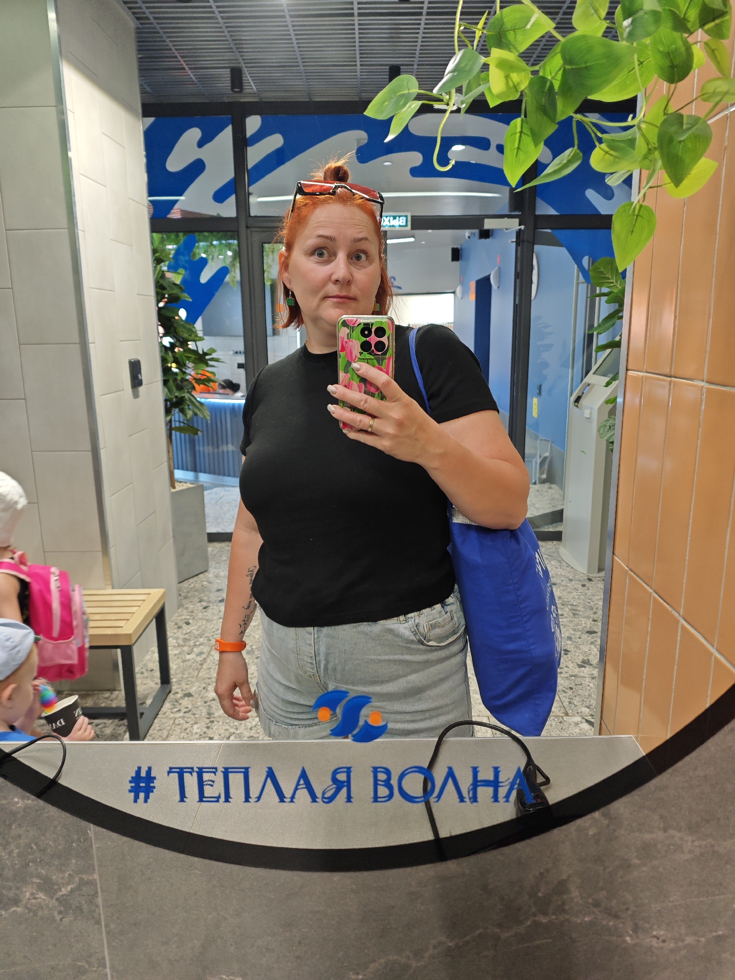 A woman stands in front of a mirror in what appears to be a recreational or commercial space. She is taking a selfie with her smartphone, which has a brightly colored case. She has reddish hair, styled in a bun, and is wearing sunglasses on top of her head. She is wearing a black t-shirt and light denim shorts. A blue tote bag hangs from her shoulder.
In the reflection, behind the woman, there is a space with blue and white decorative elements, possibly indicating a water-themed area or a swimming pool facility. A young child with a pink backpack is partially visible in the foreground, seemingly holding a treat.
The mirror has blue text, which includes the phrase "#ТЕПЛАЯ ВОЛНА," which translates to "#WARM WAVE" in English. This is likely the name or branding of the location. The setting appears to be in Votkinsk, Russia, given the Cyrillic text. Greenery is also visible near the mirror.