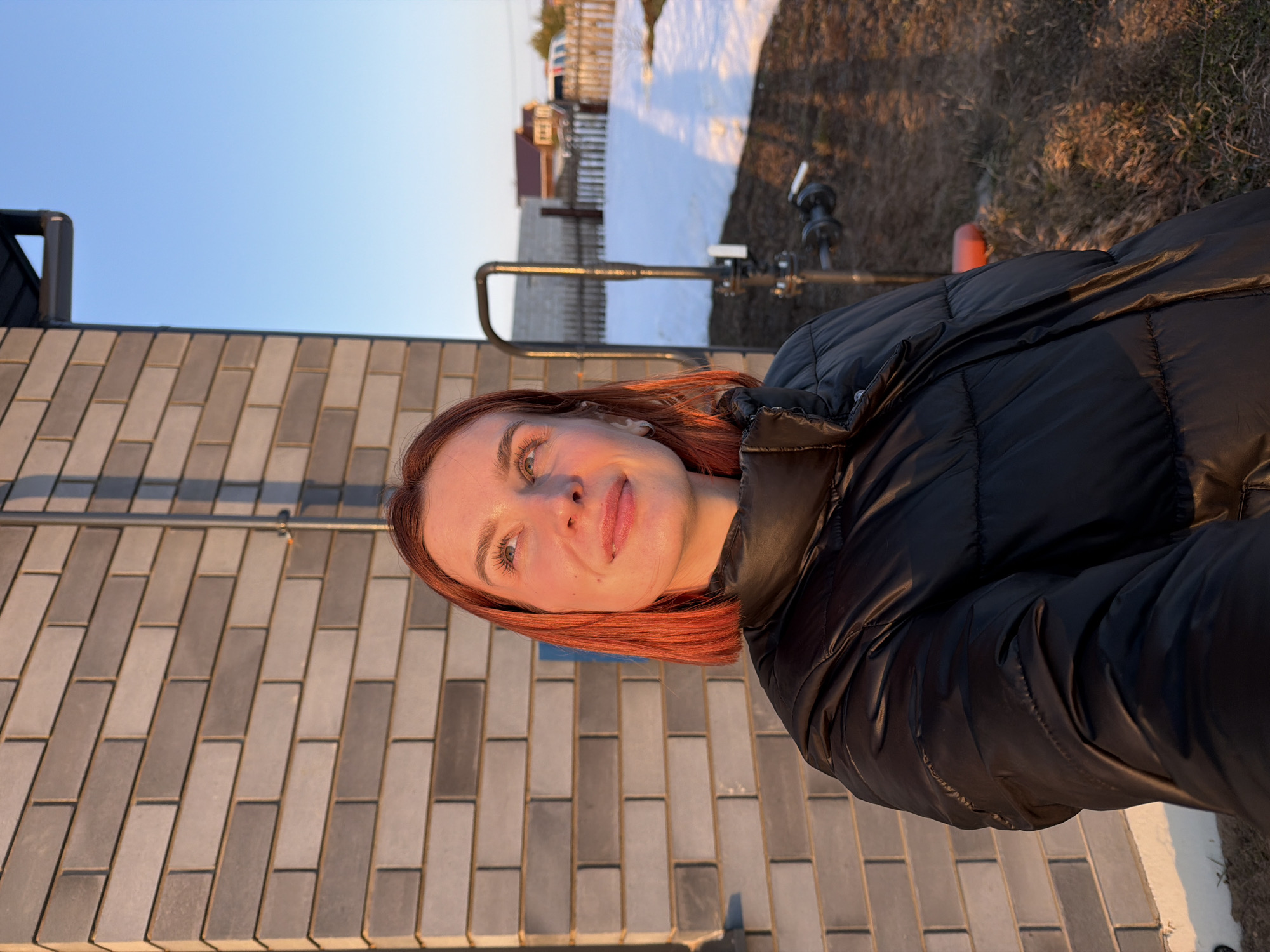 The image depicts a young adult female with short, reddish-orange hair and a light complexion, captured from the chest up in an outdoor residential setting in Chaikovsky, Russia. She wears a black quilted puffer jacket and gazes upwards and to her right with a slight smile. Her face and hair are illuminated by warm, golden light, suggesting sunrise or sunset.
To her left, a building wall constructed of light grey and darker grey bricks is visible. A thin metallic pipe runs vertically along this wall. To her right and in the background, the ground features patches of melting snow interspersed with dry, brown grass. A dark metal utility pipe or structure with visible valves stands in the mid-ground. Further beyond, a light-colored wooden fence separates the area from distant residential buildings with pitched roofs and a white vehicle. The upper right portion of the frame shows a clear, bright blue sky.