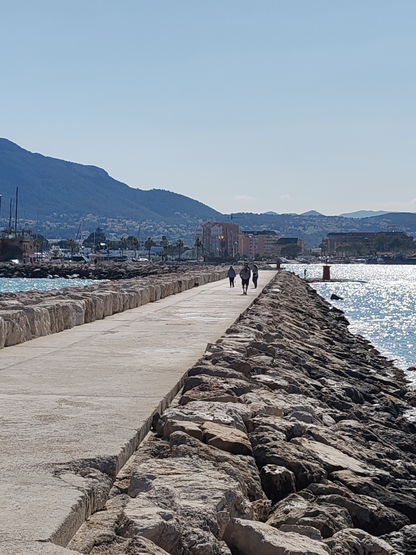 The image presents a sunlit view of the Dénia Port breakwater (also known as the Northern breakwater) in Dénia, Spain. A wide, light-grey concrete pathway, showing signs of weathering and minor surface irregularities, extends longitudinally into the water. Its left edge is formed by a vertical wall of regularly stacked, light-colored stone blocks, separating the pathway from the calmer, turquoise waters of a marina. Its right edge is defined by a sloping embankment of irregular, rough-hewn natural rocks, descending into the intensely sun-drenched open sea, which exhibits significant surface sparkle due to direct illumination.
Three individuals are walking in single file away from the camera along the center of the concrete pathway in the mid-ground. The lead person, appearing male, wears dark shorts and a dark t-shirt. Behind him, two individuals, both appearing female, wear light-colored tops and dark lower garments. They maintain a consistent spacing of approximately 1.5 to 2 meters between them.
At the distant end of the breakwater, a small, cylindrical red lighthouse with a conical top is prominent on the right side. Within the marina area to the left, multiple boats with visible masts are docked. Beyond the marina, several multi-story buildings with red-tiled roofs are visible on the distant shoreline, one displaying a Spanish national flag. The background features a large, dark-blue mountain range, identified as the Montgó Massif, dominating the left side, with lower, lighter-blue hills extending across the horizon to the right. The sky above is uniformly clear and bright blue, devoid of clouds. The scene is under bright, direct sunlight, indicating midday or early afternoon conditions.