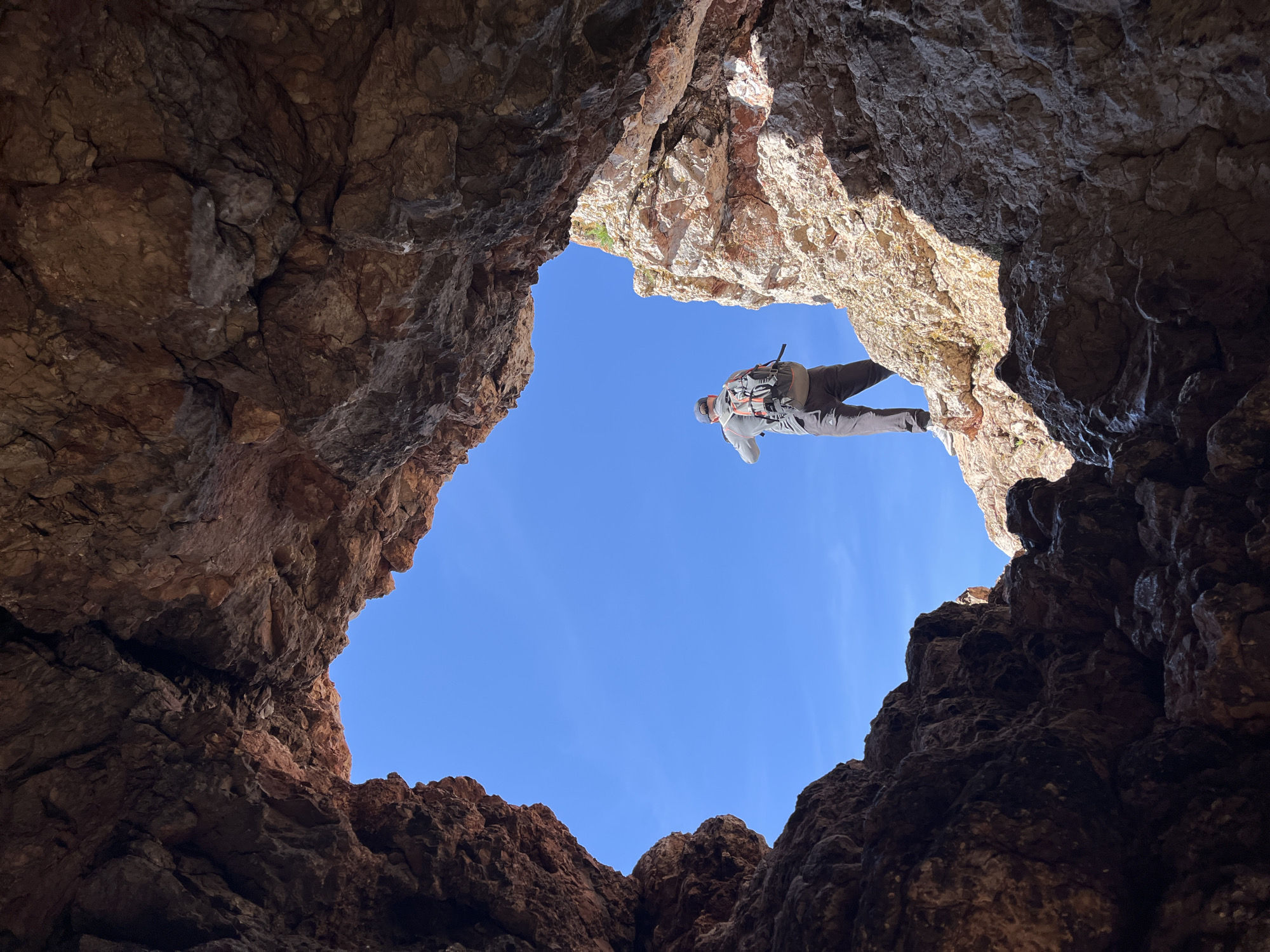A low-angle shot looking up from inside a rocky cavern reveals a hiker scaling the opening against a clear blue sky. The cavern walls are jagged and textured, predominantly in shades of brown. The hiker, wearing neutral-toned clothing and a backpack, is positioned near the upper right edge of the opening. The location is unknown.