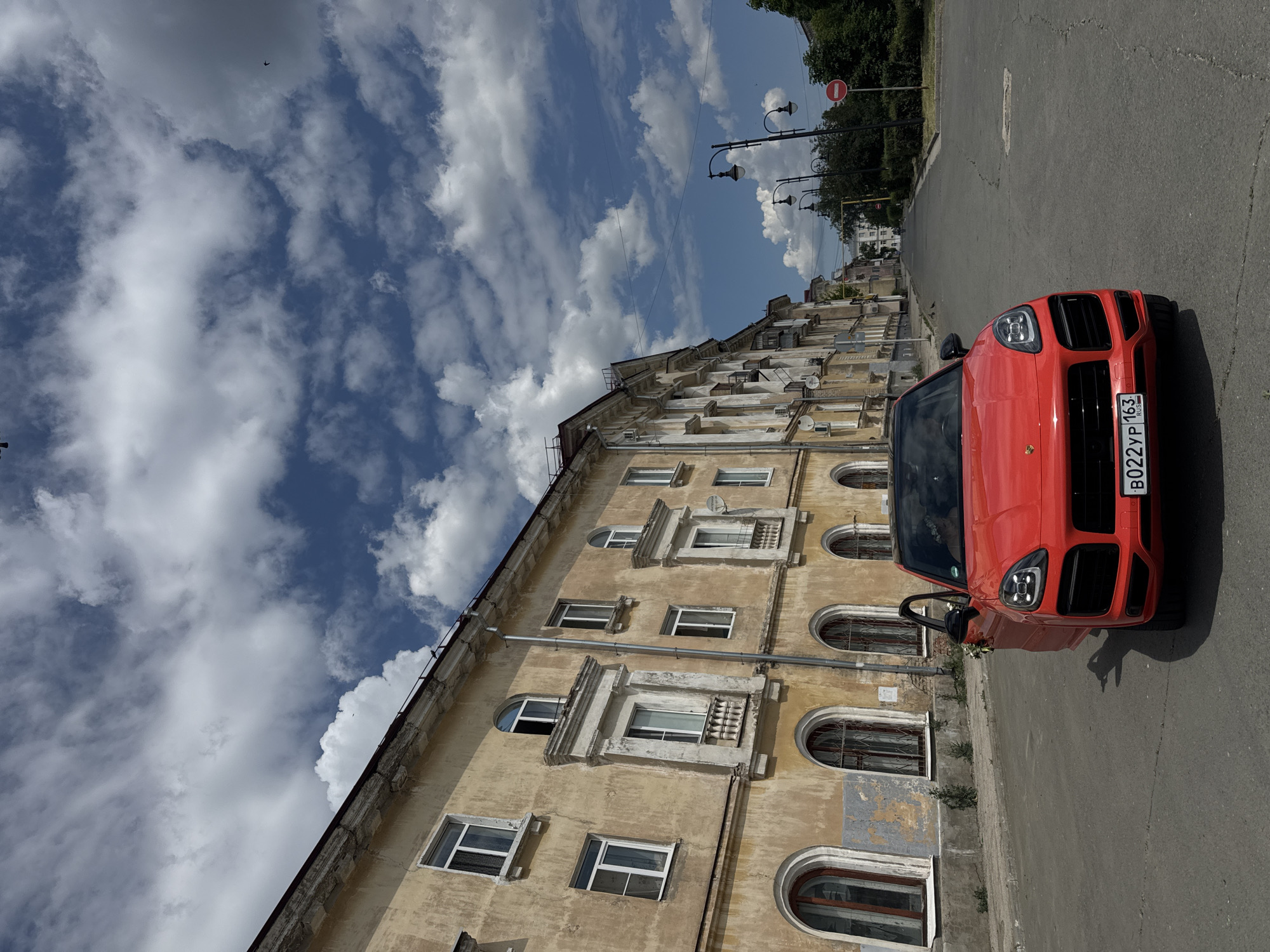 An eye-level, medium shot shows a bright red Porsche Cayenne parked on a street in an urban environment. The car is angled towards the right, with its driver's side door open, revealing white floral decorations on the interior. The license plate reads "B022YP 163 RUS".
The street is paved and appears to be a city street with a slight downward slope towards the background. On the left side of the street, a row of old, weathered, yellow buildings with ornate architectural details and arched windows stretches into the distance. The buildings show signs of age, with peeling paint and visible damage. On the right side of the street, there are more buildings, streetlights, trees, and a "no entry" sign.
The sky above is a vibrant blue with large, fluffy white clouds. The lighting suggests it is daytime, with sunlight casting shadows. The overall scene captures a juxtaposition of a modern, luxury vehicle against a backdrop of historic urban architecture under a bright, cloudy sky.